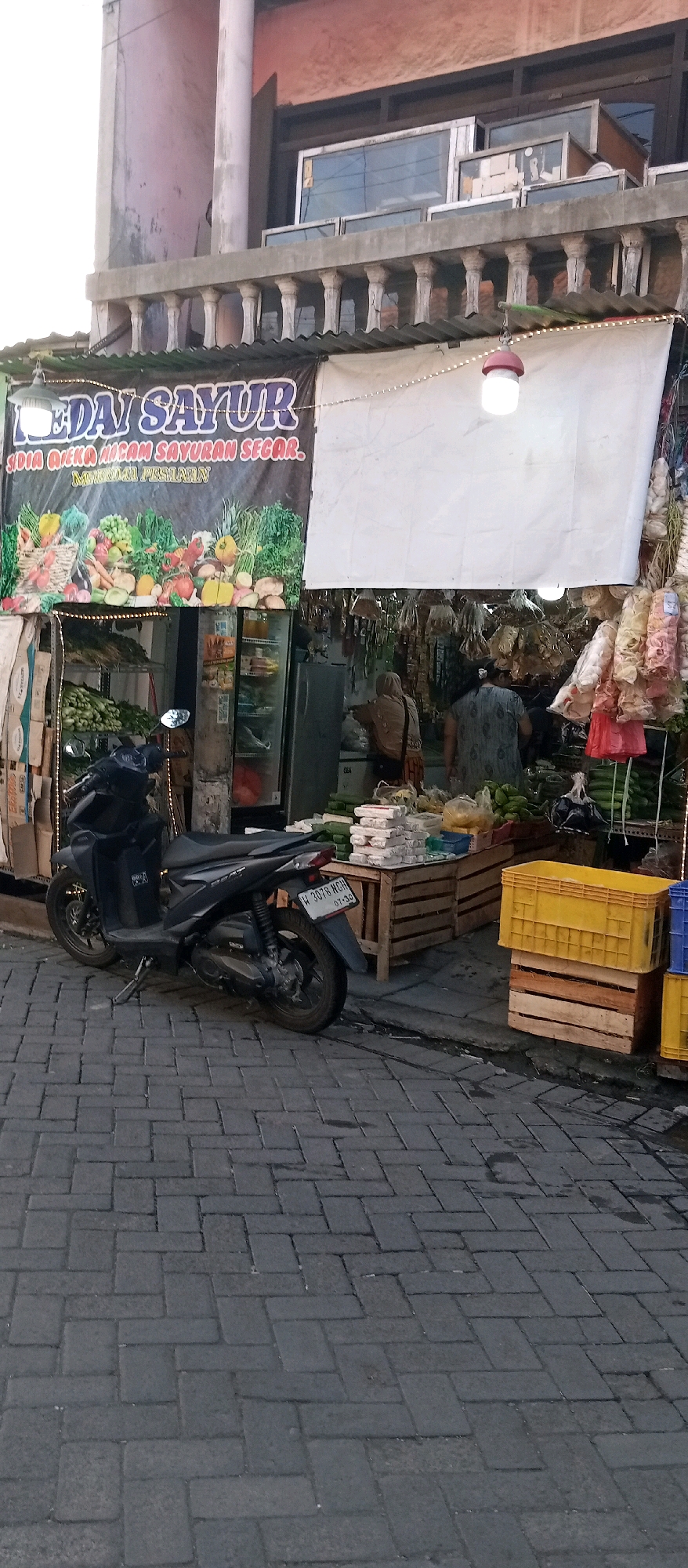 A street-level, medium shot captures a busy outdoor market stall in Wadungasri, Indonesia. A dark, modern scooter with a license plate reading "W 3078 MGH" is parked on the left, angled towards the viewer. The stall is adorned with a black banner featuring the text "KEDAI SAYUR" in purple lettering, with an illustration of various vegetables below. To the right of the banner, a blank white cloth is hung. Several vendors, including two women in headscarves, are seen interacting within the stall, attending to customers or arranging produce. The stall displays a variety of goods, including fresh vegetables, packaged snacks, and what appear to be dried goods hanging from the ceiling. In the foreground, stacked wooden crates and yellow and blue plastic crates are visible. The ground is paved with dark grey brickwork. The background reveals a multi-story building with a balcony supported by ornate pillars and windows. The lighting suggests it is daytime.