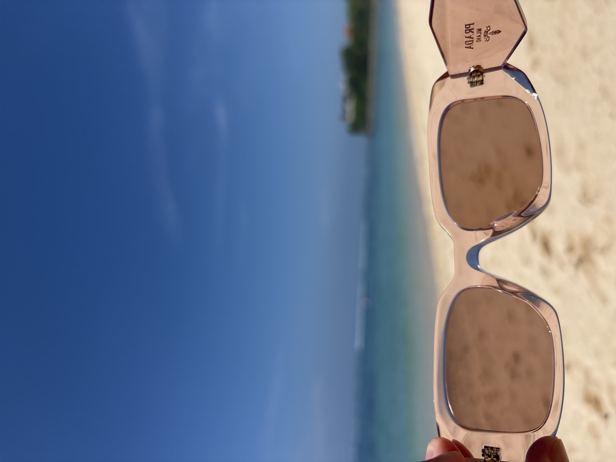 The image shows a pair of pink Prada sunglasses held in front of a beach scene. The location is Kuta Selatan, Indonesia. The sky is clear blue, and the ocean water is a gradient of blue and green. The beach is visible in the background. The sunglasses are the focal point, obstructing the view of the scene. The image is oriented vertically.