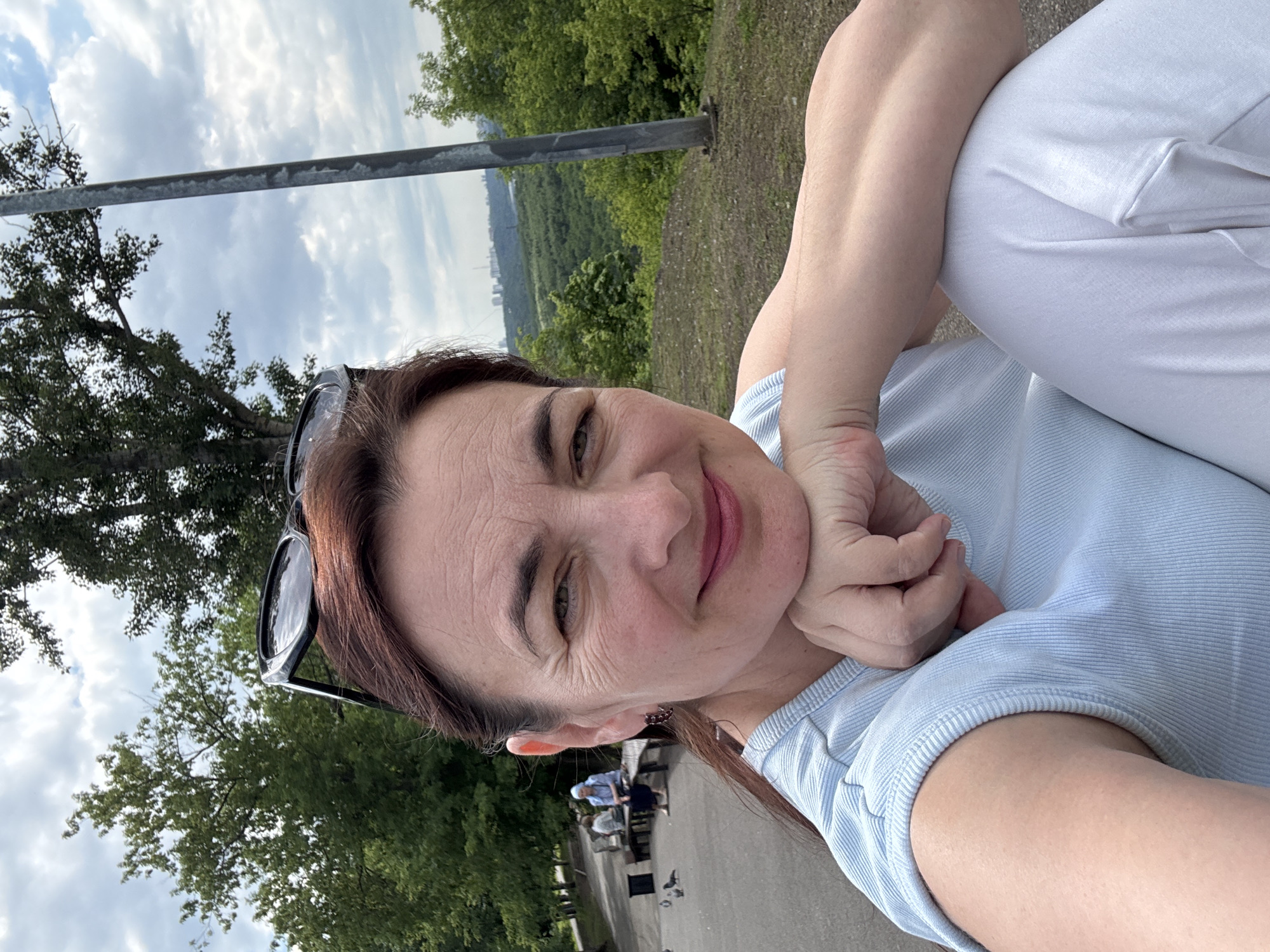 A woman with brown hair, wearing a light blue top and white pants, is taking a selfie outdoors. She has sunglasses perched on her head and is resting her chin on her hand. In the background, two people are sitting on a bench, and several pigeons are on the ground. The scene appears to be a park with trees and a cloudy sky. The cityscape in the distance suggests the location is Ufa, Russia.