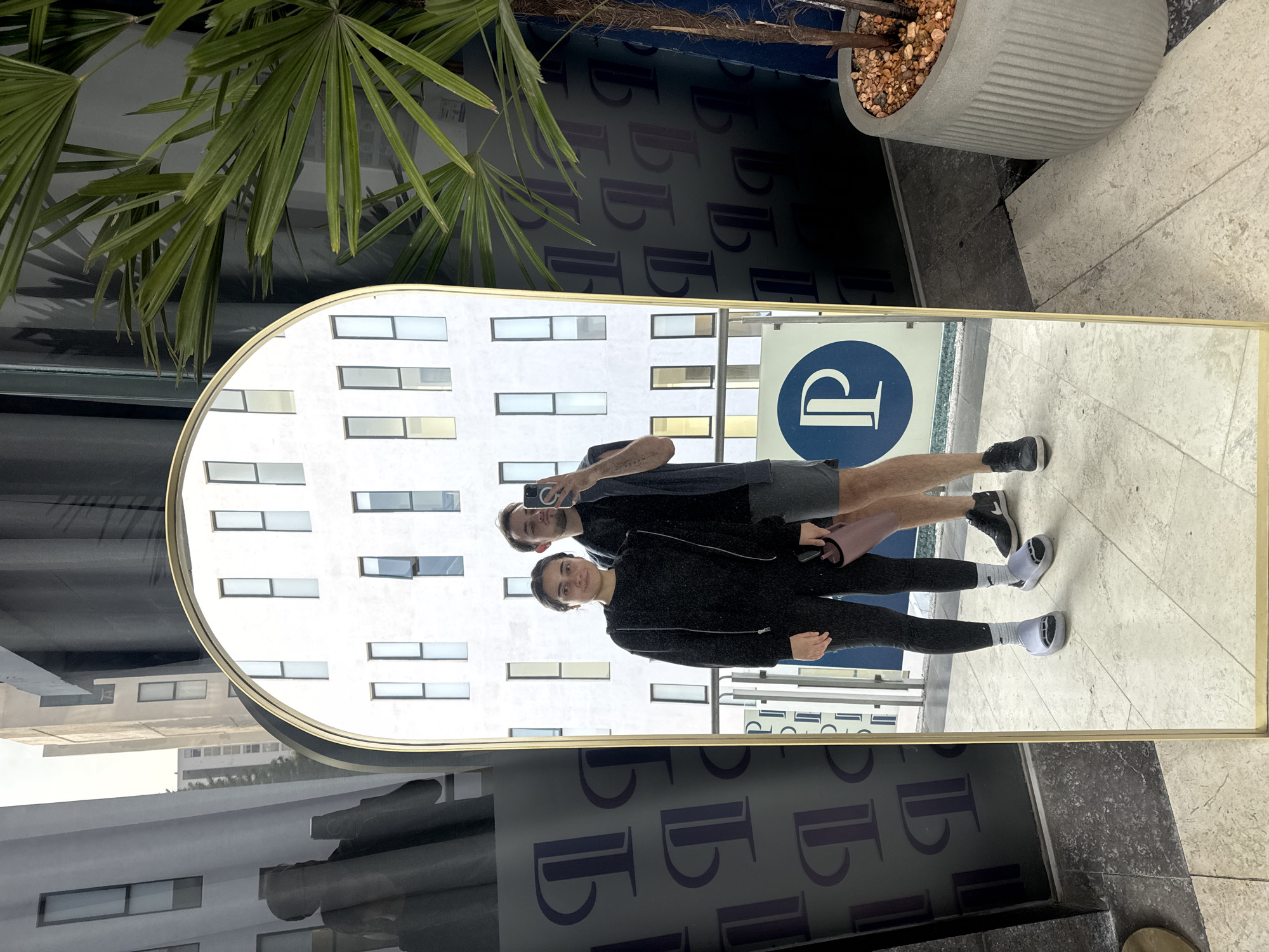 This image captures a mirror selfie featuring a man and a woman. The golden-framed mirror stands upright, reflecting a modern white building with numerous rectangular windows, indicating an urban setting. A prominent blue circular sign with a stylized white 'P' logo is also clearly reflected.
The man, on the left of the reflection, holds a smartphone up, appearing to take the photo. He wears a dark t-shirt, dark shorts, white socks, and dark sneakers. The woman, standing to his right, wears a dark zippered jacket, dark leggings, white socks, and distinct light grey, rounded shoes. She holds a light pink bag in her left hand and offers a slight smile towards the camera.
In the physical foreground and background outside the mirror's reflection, large green palm-like leaves from a potted plant are visible, along with a grey concrete planter filled with pebbles. A dark wall or surface behind the mirror features a repeating pattern of the same stylized 'P' logo seen on the sign. The bright reflections suggest it is daytime. The overall scene conveys an urban or commercial environment, aligning with the context of Puebla, Mexico.