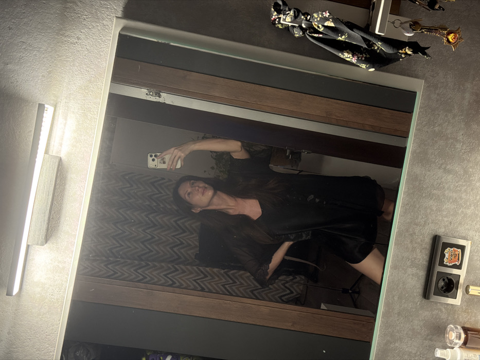 A woman in a black satin robe takes a mirror selfie. She holds her phone up and angles her body towards the reflection. The bathroom background includes a textured wall, a light fixture, a framed mirror, and various items like a scarf, keys, and toiletries scattered on the counter. The reflection shows a glimpse of a patterned bedspread or couch behind her. The scene gives an intimate, casual feel.