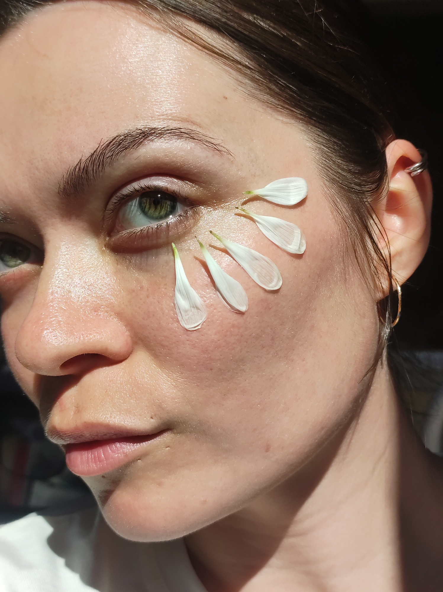 A close-up shot features a woman with fair skin and green eyes, her face partially illuminated by sunlight. Several white flower petals are placed artfully on her face, extending from below her left eye towards her temple. She is wearing a gold hoop earring in one ear and a silver earring on the upper part of the same ear. Her hair is pulled back and appears to be dark. The image is focused on the woman's face and does not show the surrounding environment, so it is impossible to ascertain if the location is Фоки, Russia or any specific city.