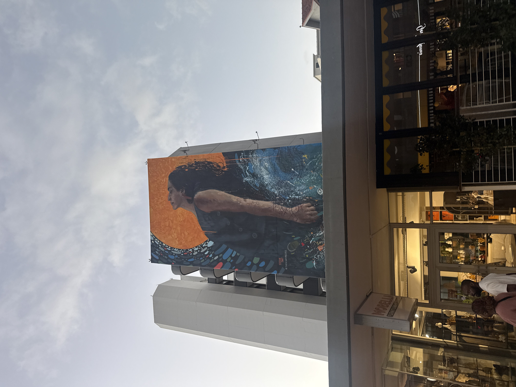 The image shows a mural of a woman swimming or floating, displayed on the side of a building. The mural has an orange background on one side and a blue water-like background on the other side. The mural is credited to "Stefaan @theses19fingers". Adjacent to the building with the mural is another building that appears to have a shop named "Morelli Furniture Lighting & Decor." People are visible inside and outside of the shop. The sky is visible above the buildings. The mural is located in Cape Town, South Africa.