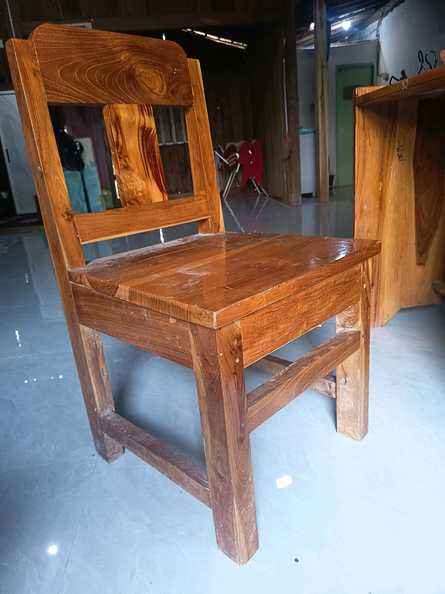 A wooden chair with a plank seat and backrest sits on a tiled floor. The chair is made of light-colored wood with visible grain patterns. A wooden table is partially visible to the right of the chair. In the background, a room with exposed beams and a corrugated metal roof is visible, suggesting an indoor or semi-enclosed space. There are some red chairs and what appears to be a white washing machine in the background. The overall environment appears to be rustic and possibly in a workshop or home setting in Sidorejo, Indonesia.