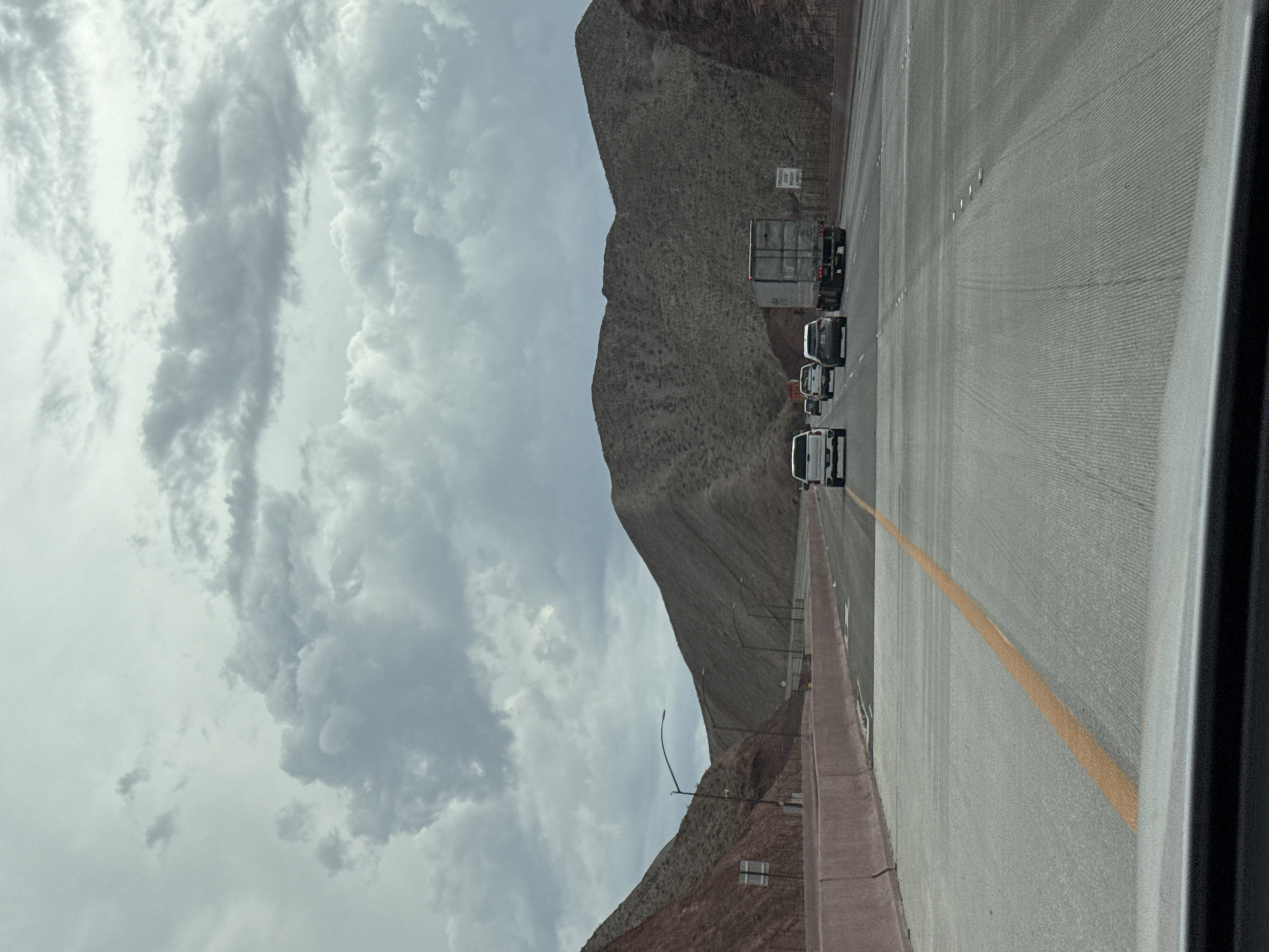 The image depicts a view from a vehicle traveling on a highway, likely Interstate 11, in Boulder City, Nevada, United States of America. The highway is flanked by arid, mountainous terrain on both sides, characterized by slopes with sparse vegetation and rocky outcrops. The sky is overcast with heavy, dark gray clouds, suggesting a potentially stormy or gloomy day.
Several vehicles are visible on the highway, including a white pickup truck in the right lane, a silver SUV and a white sedan in the left lane ahead of the viewpoint, and a large gray semi-trailer truck in the right lane further ahead. The road surface is concrete with visible lane markings, including a solid yellow line on the left and intermittent white lines delineating lanes. Concrete barriers line the sides of the highway. Streetlights are present along the route.
The scene is devoid of any identifiable people. The overall impression is one of travel through a desolate, natural landscape under a somber sky.