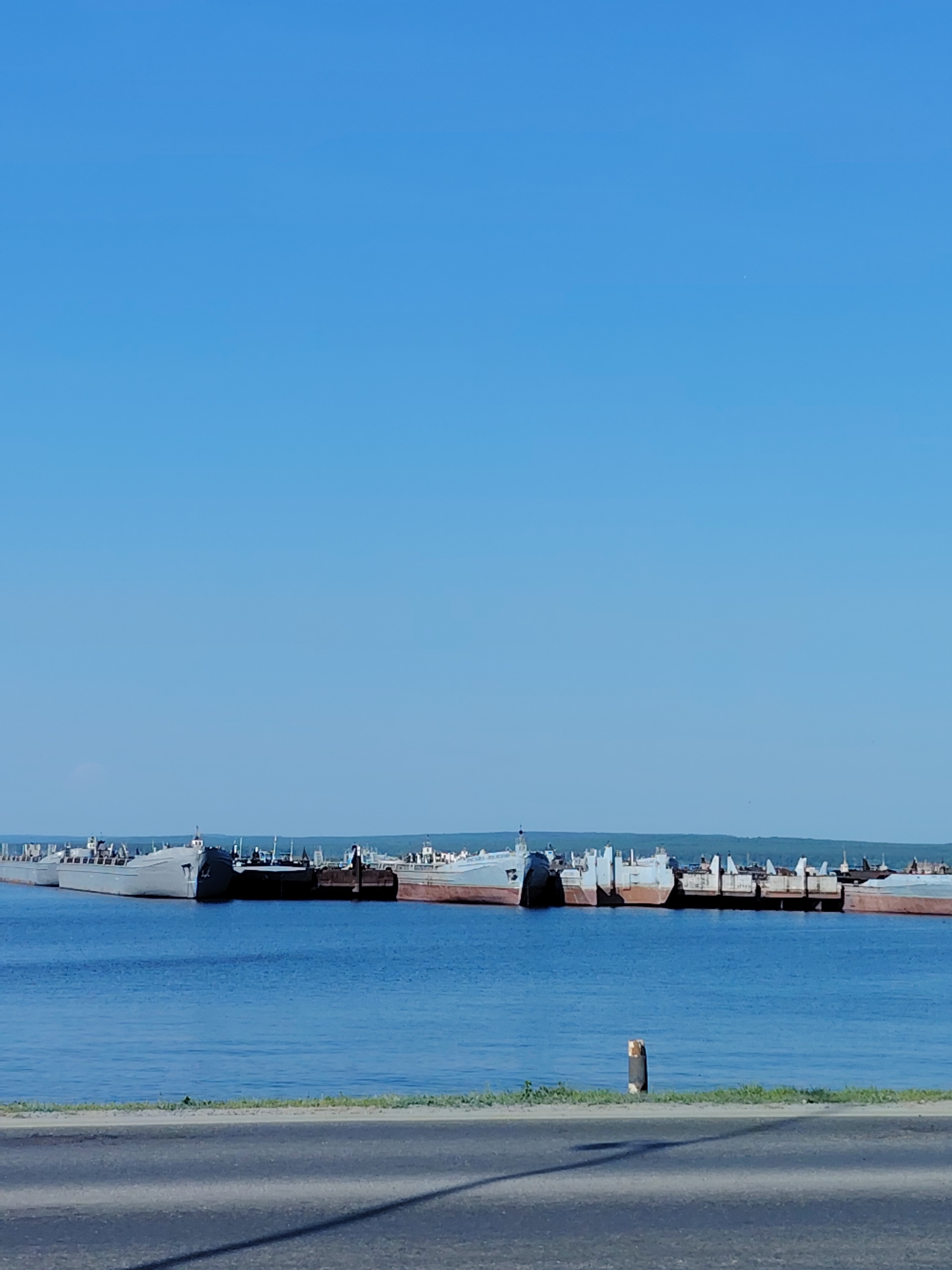 The image depicts a row of barges docked in a body of water, likely a river or reservoir, in the city of Chaikovsky, Russia. The sky is clear and blue. A paved road runs along the foreground, separated from the water's edge by a strip of vegetation. A wooden post is visible near the edge of the water. The barges are various shades of gray and brown and appear to be tied together. The opposite bank of the water features a line of trees.
