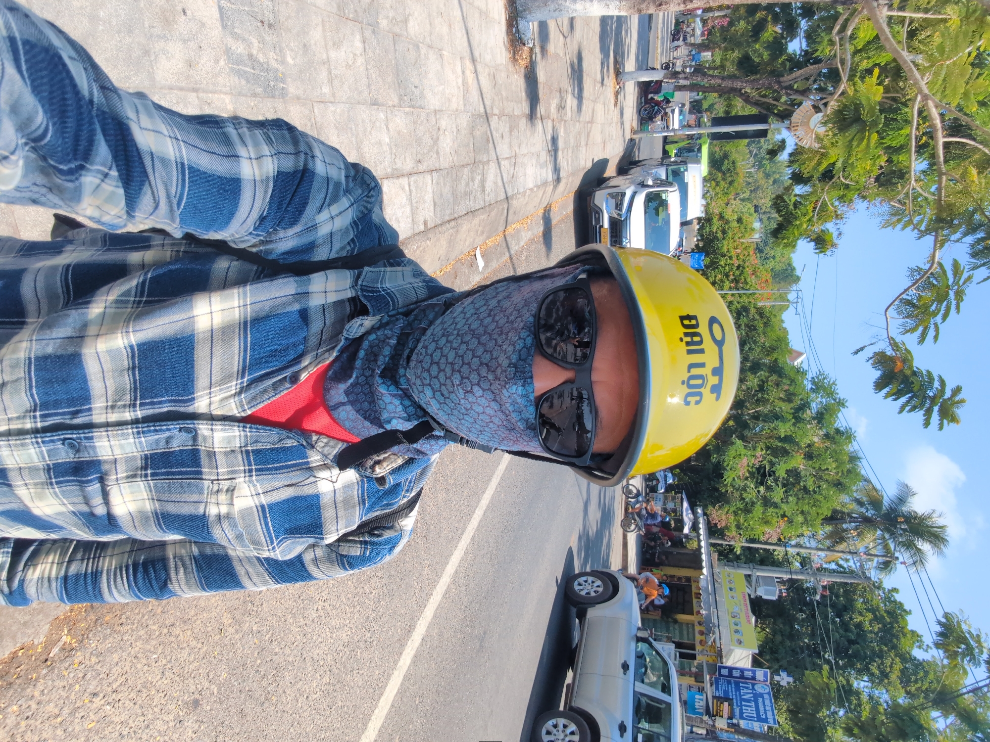 A man wearing a blue and white plaid shirt over a red undershirt, a patterned blue and gray face covering, sunglasses, and a yellow helmet stands on a street in Phú Quốc, Vietnam. The helmet has the Vietnamese words "Đại Lộc" printed on it. He appears to be taking a selfie. Behind him, a white truck is parked along the side of the road. Further down the road, other vehicles and people can be seen. Shops and trees line the street, and the sky is blue.
