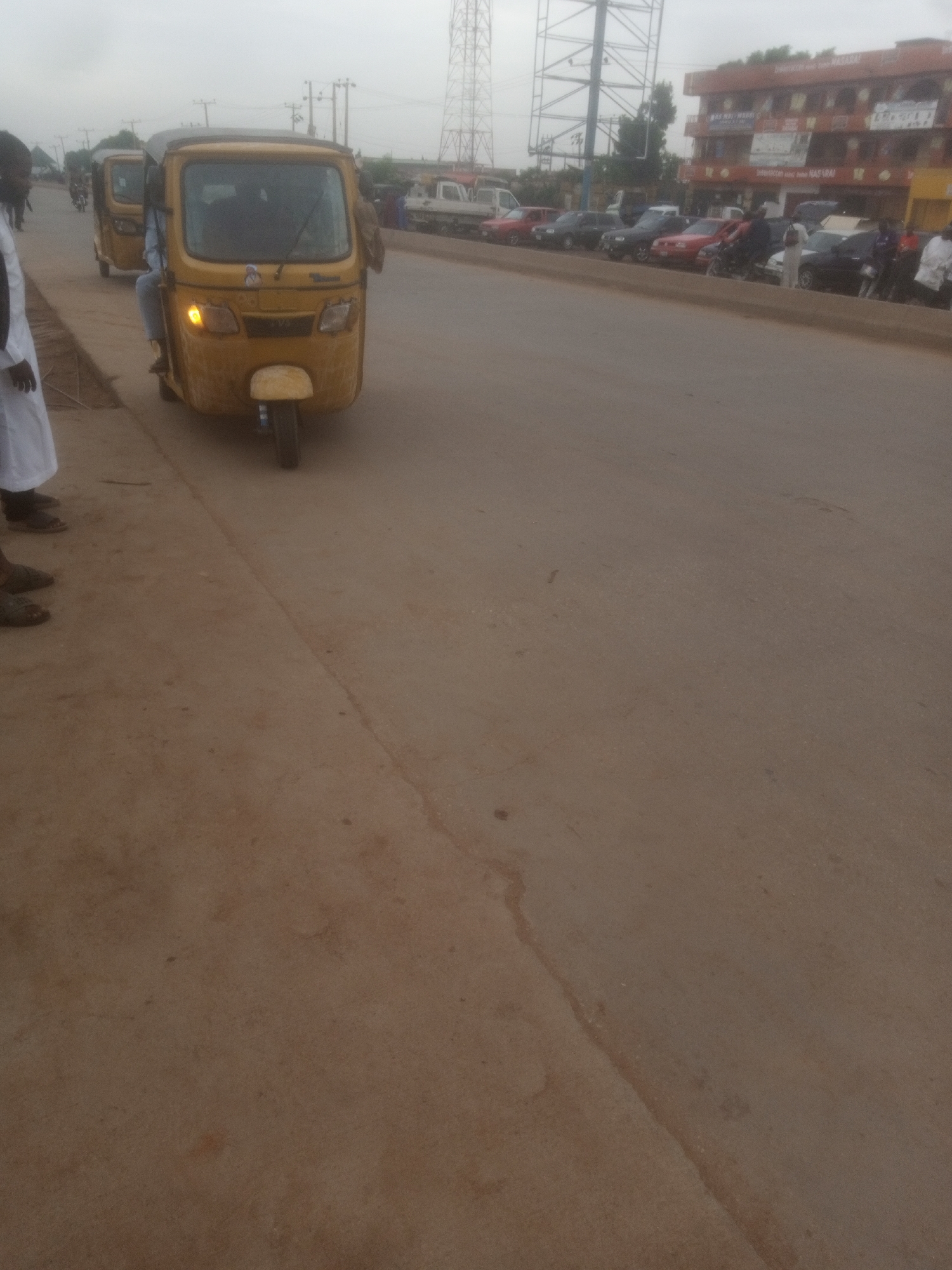 In Tundun Wada South, Nigeria, a yellow auto-rickshaw is parked on a dirt road next to a paved road. The auto-rickshaw is facing left and has its headlights on. A person wearing a white robe and sandals is standing in the foreground to the left of the auto-rickshaw. Several other auto-rickshaws are visible in the background, as well as other vehicles and buildings. The sky is overcast.