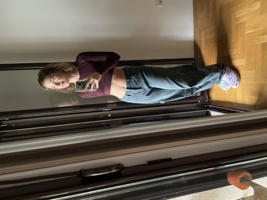 An indoor scene in Boreti, Montenegro, features a female subject standing in front of a large reflective surface, likely a mirror or glass door, taking a self-portrait with a smartphone held in her right hand. The subject has light brown, curly hair and is looking upwards and to her right. She is dressed in a long-sleeved, cropped maroon top, exposing her midriff and a tattoo below her navel, paired with loose-fitting teal-grey trousers. On her feet are light purple Crocs adorned with colorful Jibbitz. The flooring is light-toned wooden parquet. To the subject's right, a plain white wall is visible with a single white electrical outlet. To her left, a dark-framed window or glass door extends vertically, and a partial view of an orange skateboard wheel is visible at the lower left, resting near the frame. The specific location within Boreti is not discernible from the interior details.