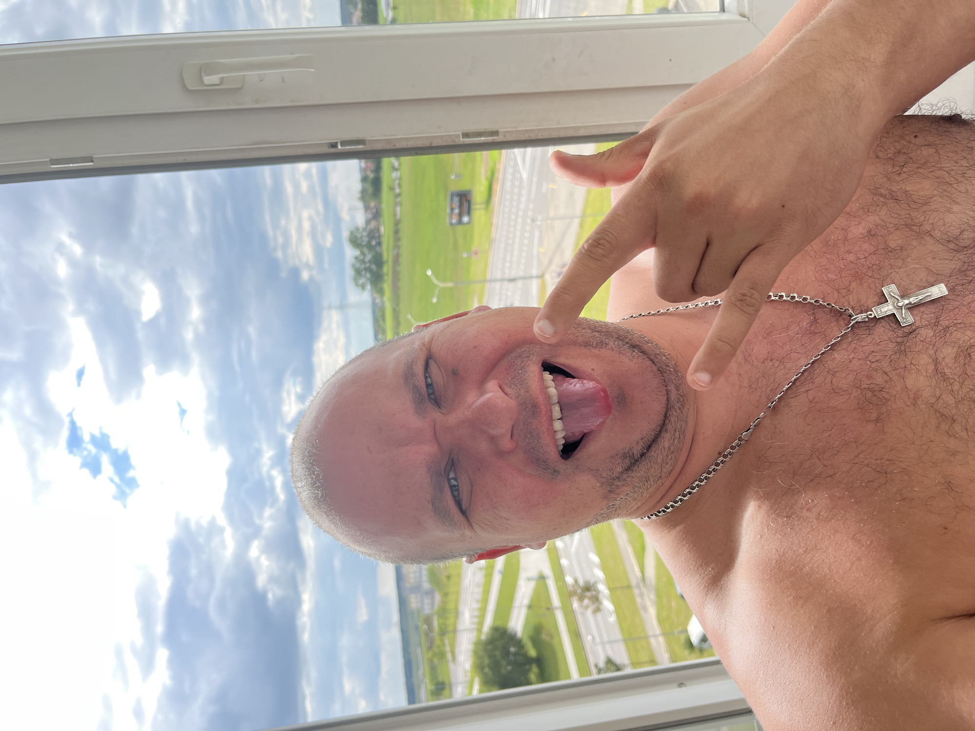 A man, shirtless and wearing a silver chain with a cross pendant, poses for a selfie by a window. He has a shaved head and is sticking his tongue out, making a "rock on" gesture with his fingers. The background shows a cloudy sky and a cityscape that appears to be Minsk, Belarus, with roads, buildings, and trees visible.