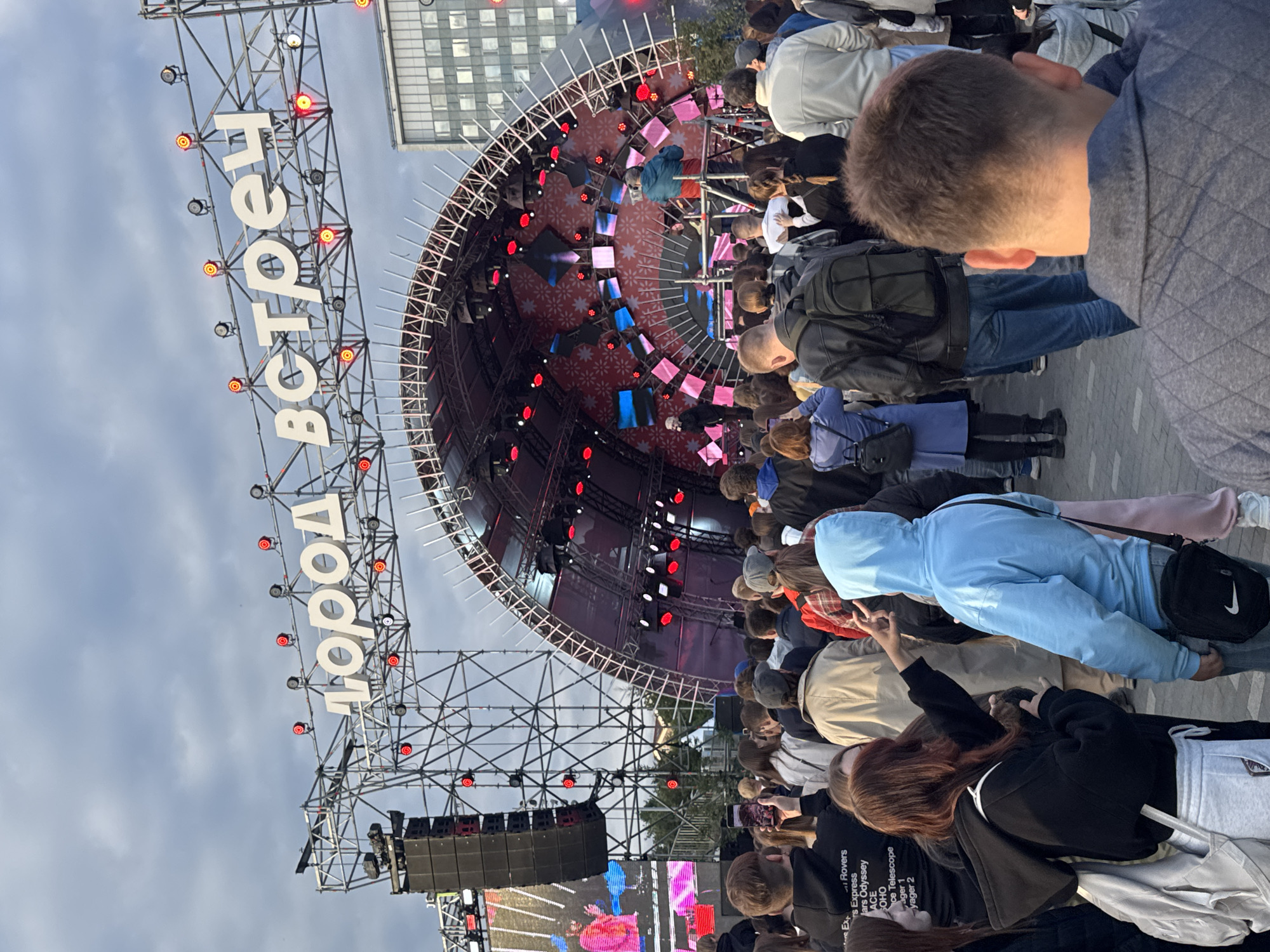 This image depicts an outdoor event with a large crowd gathered in front of a stage. A prominent sign above the stage displays white Cyrillic lettering that reads "ГОРОД ВСТРЕЧ" which translates to "City of Meetings." The stage itself is a large, arched structure with a colorful backdrop featuring geometric patterns and illuminated panels. Several individuals are visible on the stage, including a performer and what appear to be technical crew members.
The foreground shows a dense crowd of people facing the stage, with many holding up their phones to take pictures or record videos. The individuals are dressed in casual clothing, and the atmosphere suggests a concert or festival. The event appears to be taking place in a public square or open area, with buildings visible in the background, characteristic of an urban environment. The sky is overcast, indicating either early morning or late afternoon.
This event, "City of Meetings," is an annual festival that takes place in the city of Perm, Russia, in the central square.