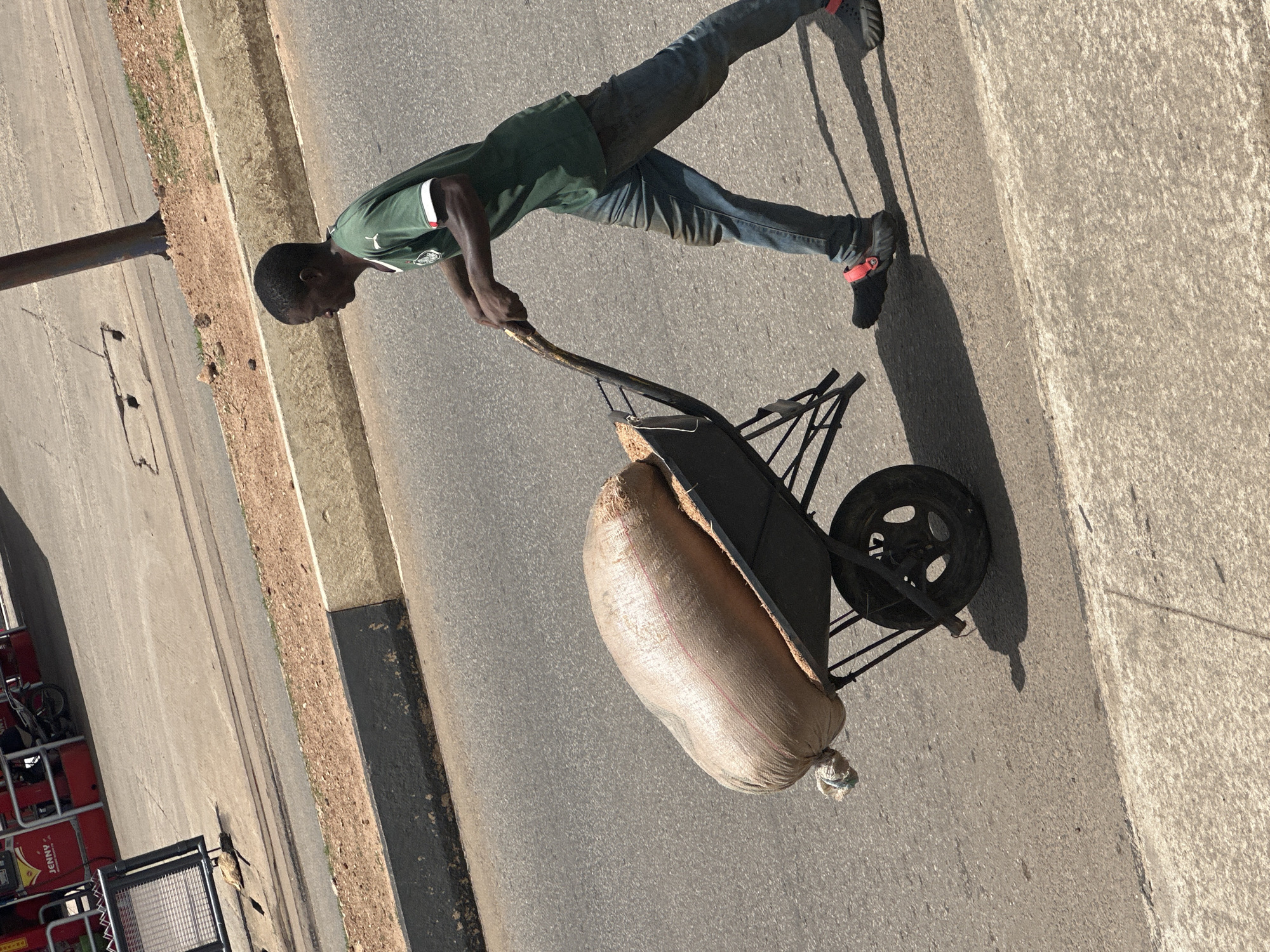 A man is pushing a wheelbarrow filled with a large sack of grain down a paved street in Gura Top, Nigeria. The man is wearing a green t-shirt and blue jeans. The sack is made of woven material and appears to be tightly packed. The wheelbarrow has a single wheel and two handles, which the man is gripping. The street is asphalt and appears to be in good condition. There is a sidewalk to the left of the street, and a grassy embankment beyond that. In the background, there is a red building with the word "JENNY" visible on it, suggesting a commercial establishment. The sun is shining brightly, casting a shadow from the man and the wheelbarrow on the ground.