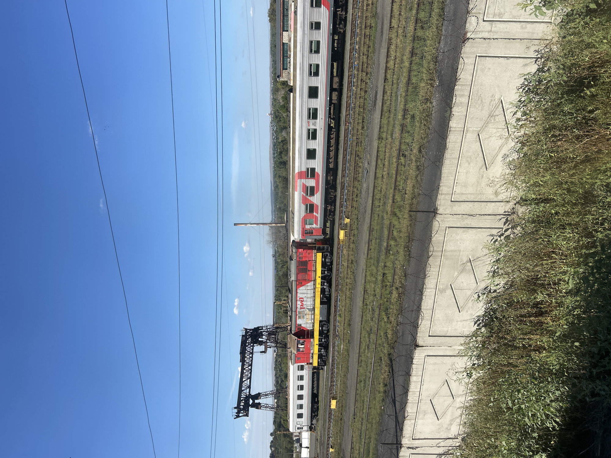 The image depicts a train yard in Orsk, Russia, under a clear blue sky with scattered clouds. In the foreground, a concrete wall with diamond-shaped indentations and barbed wire on top separates the viewer from the train tracks. Tall, dry grass grows in front of the wall. Several sets of train tracks stretch across the scene, with some areas of sparse green grass and darker, rougher ground.
In the middle ground, a train is parked on the tracks. The engine is red and yellow with Russian text and logos. It is coupled to several passenger cars that are silver with red stripe detailing and the same Russian text. To the left of the train, a large, industrial gantry crane stands silhouetted against the sky. Further in the background, more train cars, buildings, trees, and hills are visible. A tall industrial chimney or smokestack is also present in the distance. The overall impression is of a functional, industrial area.