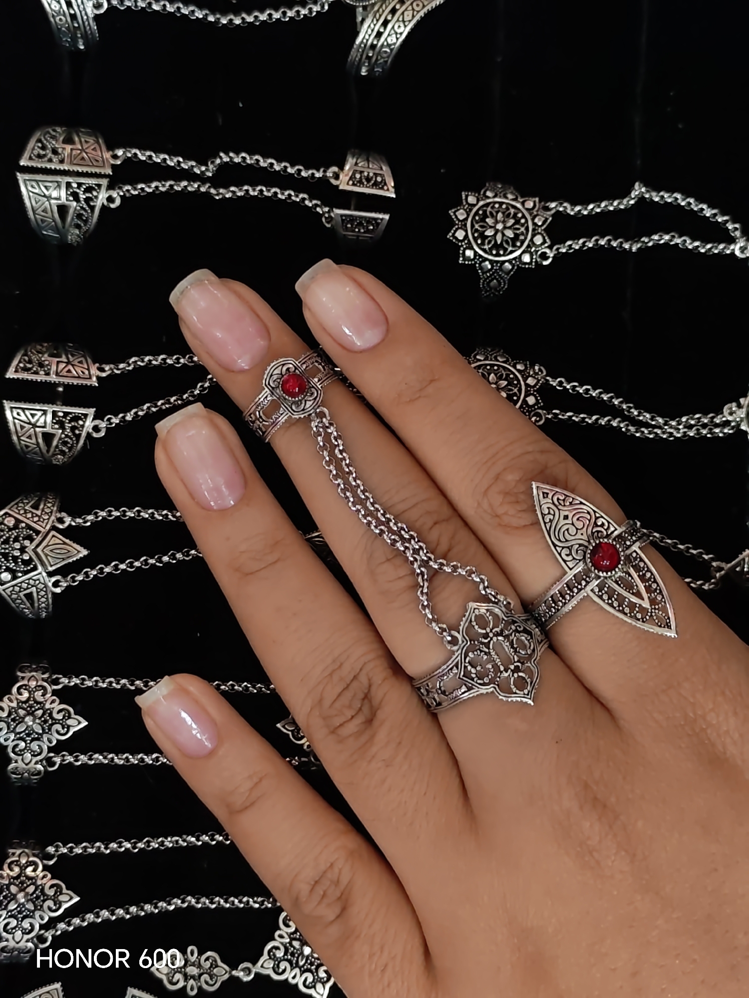 The image shows a close-up view of a person's hand wearing ornate silver rings connected by chains, against a dark, possibly black, fabric background. The rings are intricately designed with geometric patterns and small red stones. The person's fingernails are manicured and painted with a neutral polish. The background appears to be a collection of similar jewelry items, also made of silver and featuring detailed designs. There is no discernible connection to the city of Ussurijsk, Russia, in the visual information provided. The scene focuses solely on the jewelry and the hand.