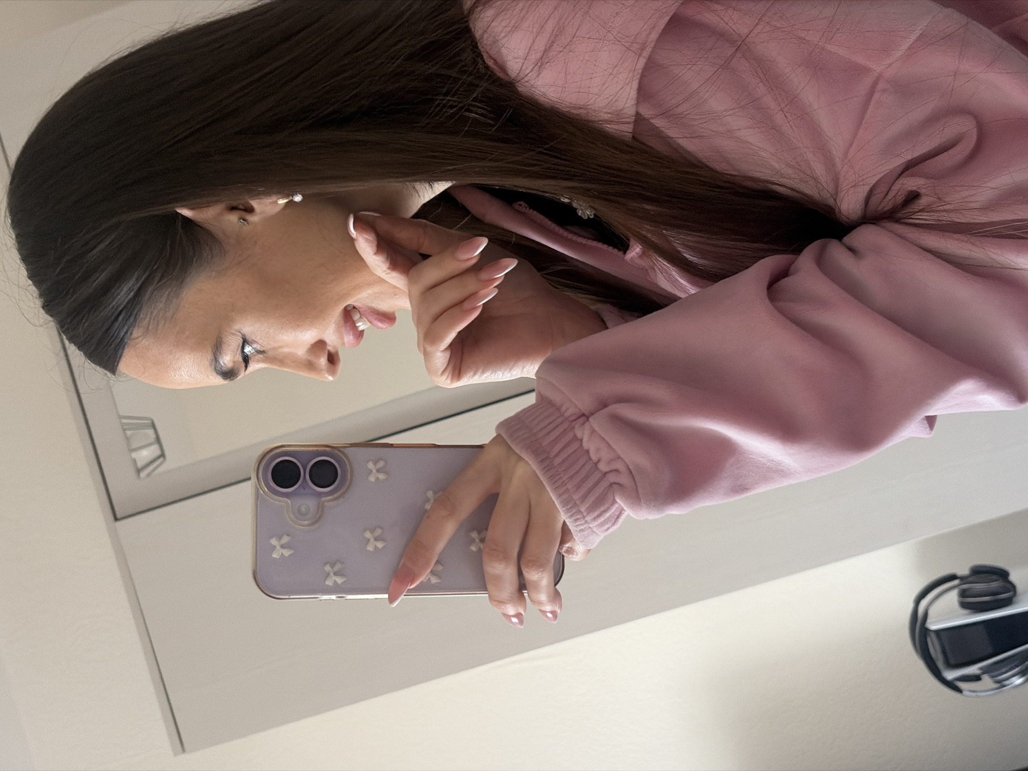 The image depicts a young adult female captured in a mirror selfie, seen from the chest up in profile, looking to her left. She has long, dark brown hair, winged eyeliner, and a faint smile with her mouth slightly open. Her right ear features a pearl drop earring and a small stud. Her nails are long, manicured with a pink base and white French tips. She is wearing a light pink, possibly velour or fleece, hooded sweatshirt with ribbed cuffs.
She holds a light purple smartphone in her right hand, equipped with a matching case adorned with small white bow embellishments and a dual-lens camera system. Her left hand is positioned near her chin. In the bottom left of the reflected background, a pair of black over-ear headphones rests on a silver stand. The reflected environment consists of plain, light-colored walls and a door frame, indicating an indoor setting. There are no discernible visual elements in the image to indicate a specific city or geographical location such as Saint Petersburg, Russia.