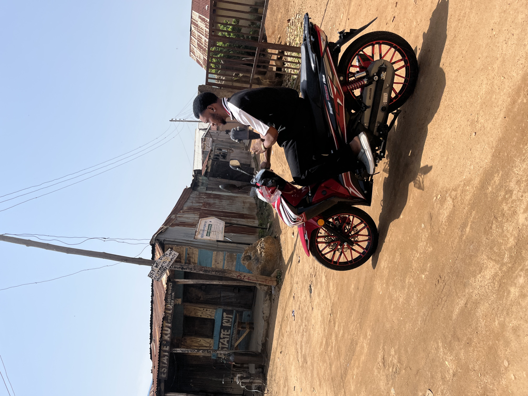 In a dusty, unpaved street in Jos, Nigeria, a young man sits astride a red and black motorcycle. He is dressed in a black t-shirt with white and red trim and black pants. His hair is short and dark, and he sports a beard. The man is looking to his left, his hands on the handlebars. The motorcycle has red spoked wheels and the words "Super Hunter Plus" are visible on its side.
Behind the man, a row of corrugated metal and wooden structures line the street. One building has the words "ESTATE KUT" and "CD" painted on its facade, along with a phone number. Another sign reads "BEN WELDING CO. STRUCTU..." Another structure has a white sign with blue text. A metal gate stands to the right of the man. The sky is clear and blue. The overall impression is one of a vibrant, informal urban environment.