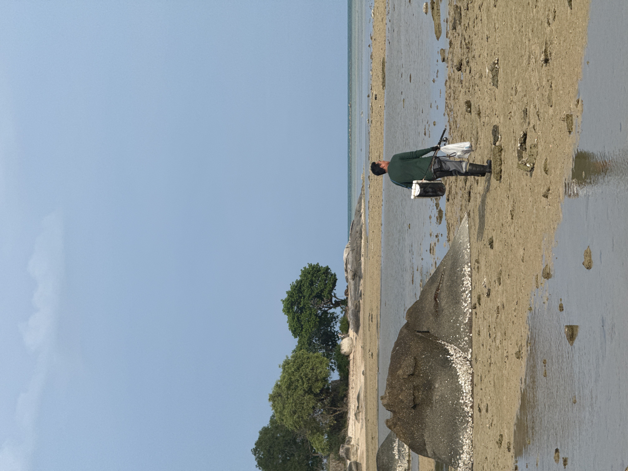 A man walks on a sandy beach with scattered rocks and shallow water. He is wearing a green long-sleeved shirt, dark pants, and black boots. He carries a white and black bucket bag over his left shoulder and a fishing rod in his right hand, with a white plastic bag hanging from it. The beach is lined with trees and rocks, and the sea stretches to the horizon under a clear blue sky with a few wispy clouds. The location is Baan Harn, Thailand.