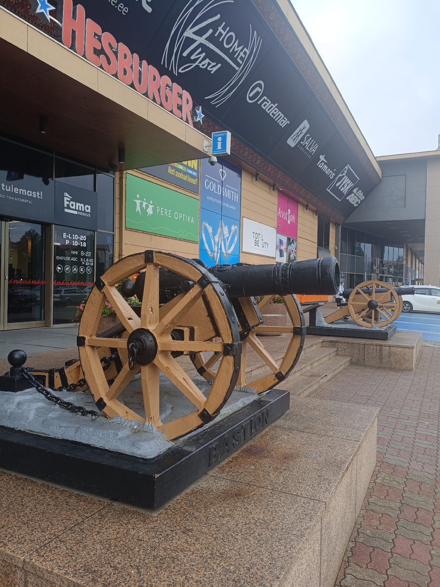 In Narva, Estonia, two decorative cannons with large wooden wheels are positioned on a stone platform in front of a modern commercial building. The building features large glass doors and windows, with signs for businesses such as "Hesburger," "Fama Keskus," "Pere Optika," and others visible. The cannons are connected by thick chains to a grey base. The ground in front of the platform is paved with red and grey interlocking bricks. A white car is parked on the right side of the image. The sky is overcast.