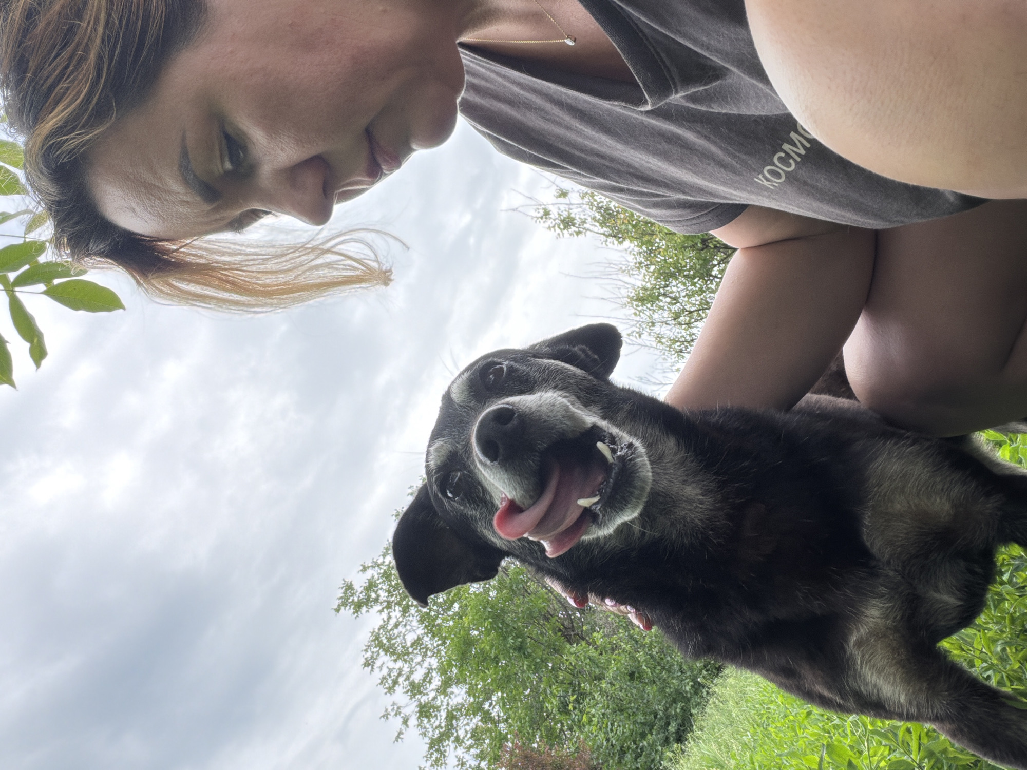 A woman and a dog are in a field near Leningradskaya, Russia. The woman kneels and looks down at the dog, who is black with graying fur around the muzzle. The dog is sticking its tongue out. The sky is overcast with some greenery visible. The woman is wearing a gray t-shirt with white lettering. The ground is covered in green vegetation.