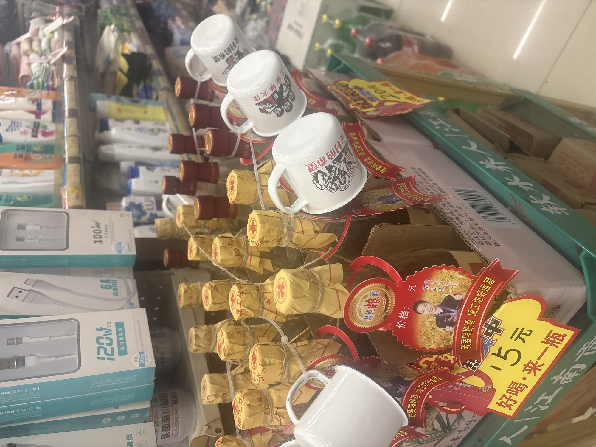The image shows a close-up of a shelf in a store, likely a supermarket or convenience store, in Suzhou Industrial Park, China. The focus is on a display of small bottles of alcohol, each individually wrapped in yellow paper and tied with twine. Several white ceramic mugs with red Chinese characters printed on them are placed in front of the bottles, some of them are attached to the bottles. One of the mugs appears to have a depiction of people on it. A large, colorful price tag is prominently displayed, indicating a price of "15元" (15 Yuan) and the text "好喝·来一瓶" (delicious, buy a bottle). Another red ribbon banner reads "我要喝好酒 顺工坊好酒造" (I want to drink good wine, Shun Gong Fang makes good wine). Behind the alcohol display, various other products are visible, including what appears to be mobile phone charging cables in branded packaging on the left, and bottles of beverages and toiletries on shelves in the background. The overall environment suggests a retail setting with goods neatly arranged for sale.