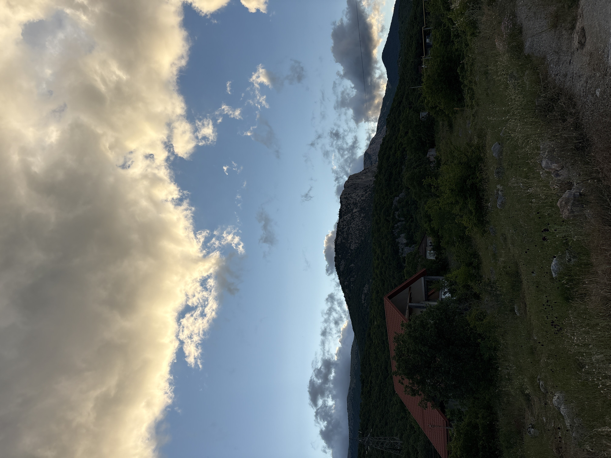 The image depicts a rural landscape with a dramatic sky. In the foreground, a dirt path winds through dry grass and scattered rocks. A house with a prominent orange-tiled roof is situated on the left side of the frame, surrounded by dense green trees. Behind the house, the terrain rises into a series of lush, tree-covered hills. The background is dominated by a large, imposing mountain with a rocky, lighter-colored peak. The sky is a mix of clear blue and voluminous clouds, some of which are illuminated with a warm, golden light, suggesting either sunrise or sunset. A single, tall, metal transmission tower is visible on the far left, partially obscured by trees. The scene appears to be in a relatively undeveloped, mountainous region.