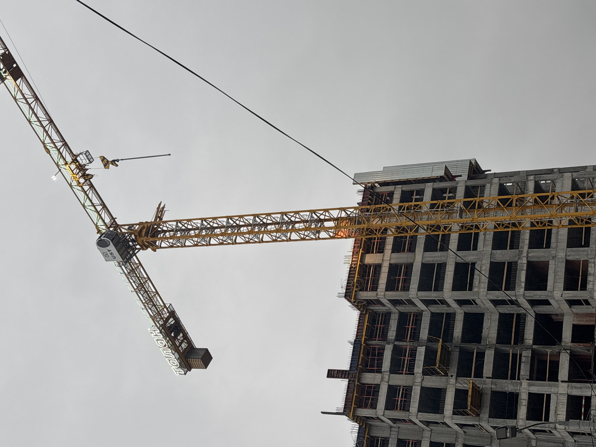 A low-angle shot of a construction site in Perm, Russia, depicts a high-rise building under construction adjacent to a tower crane under gray sky. The building's concrete structure is visible. The crane has a visible arm extending horizontally towards the building, with "ПОДЪЕМ" printed vertically on its side. A cable stretches across the sky, intersecting the building's highest point.
