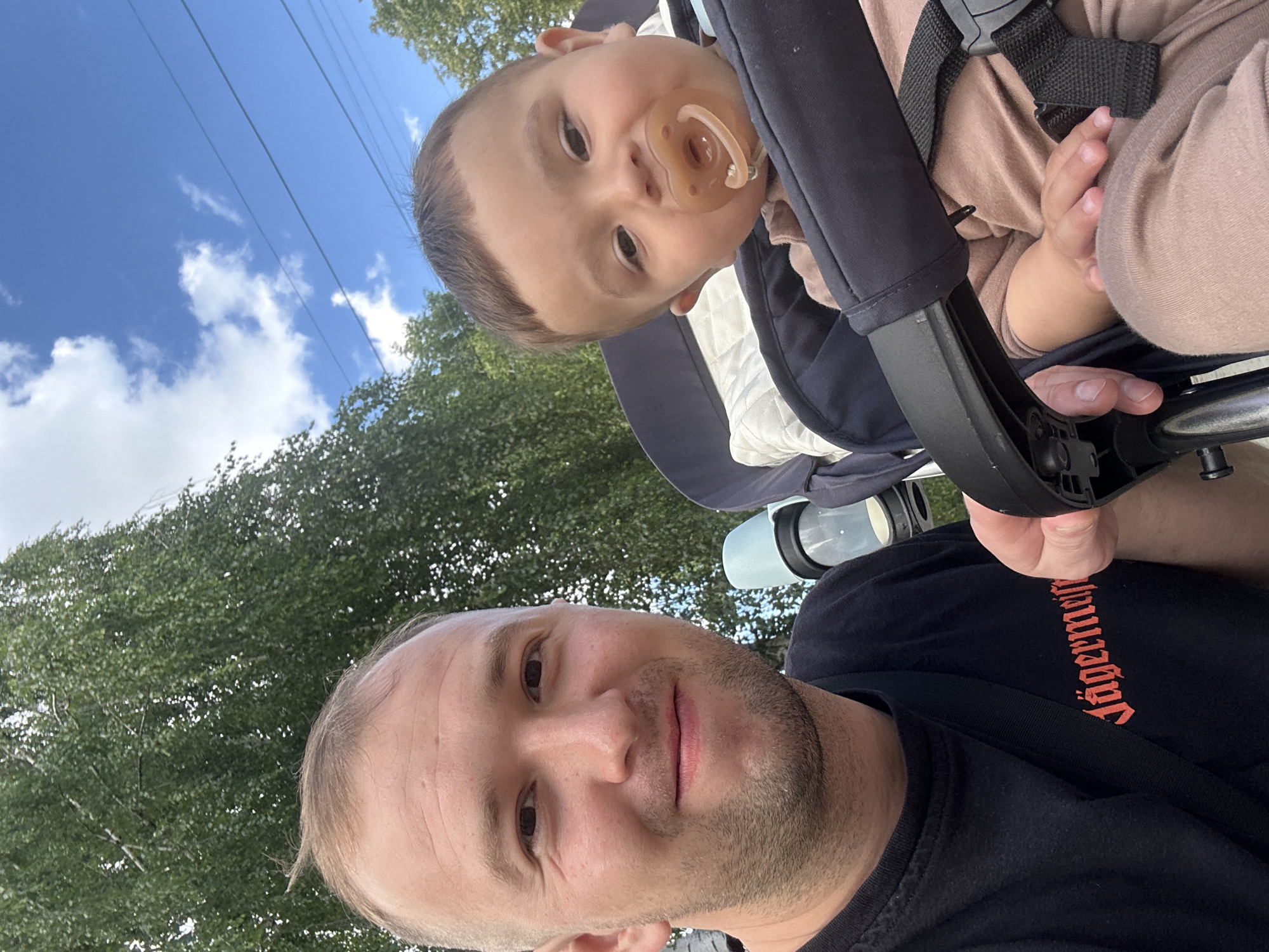 A man and a baby are outdoors in a stroller. The man is in the foreground on the left, smiling and looking at the camera. He is wearing a black t-shirt with "Jägermeister" printed in red gothic font on it. The baby is in a stroller to the right of the man, also looking towards the camera with a pacifier in its mouth. The baby is wearing a light brown outfit. The background consists of green trees and a blue sky with white clouds. There is a water bottle holder attached to the stroller. The image appears to be taken during the daytime. The scene is set in Chaikovsky, Russia, as indicated by the specific location information provided.