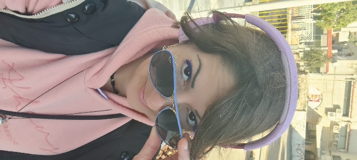 A close-up, selfie-style portrait depicts an adult female outdoors in Nahariyya, Israel, under daylight conditions. The subject is positioned centrally, looking directly at the viewer with a slight smile. She has dark brown hair and is wearing light purple over-ear headphones on her head. Aviator-style sunglasses with light blue frames and dark, branded Ray-Ban lenses cover her eyes, with her right hand (viewer's left) holding the right earpiece to slightly lower them. Visible makeup includes blue eyeliner on the lower lash line and gold glitter on the outer corner of her right eye (viewer's left). She is dressed in a pink hooded sweatshirt, which appears to have faint embroidery on the chest, layered under a black zippered jacket. A black beaded choker and a long black cord necklace with a silver-colored pendant are also visible. The background features light-colored, multi-story urban buildings with mounted external air conditioning units, interspersed with green trees and bushes. A yellow metal railing is partially visible in the mid-ground. The overall environment suggests an urban street or pedestrian area.