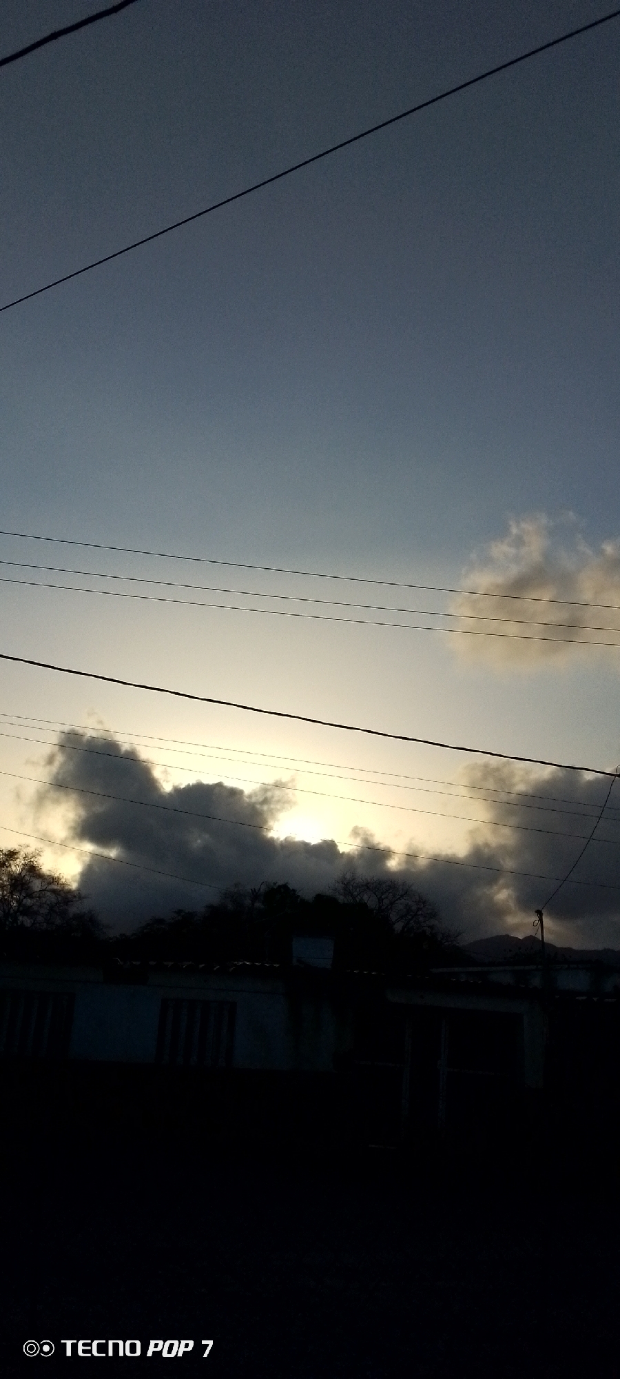 The image shows a sky with dark clouds obscuring the sun in Altagracia, Venezuela. Overhead power lines are visible. Below the sky is a dark silhouette of trees and a building. The bottom of the image is very dark, obscuring any details on the ground. The name of the camera is visible in the bottom left corner.