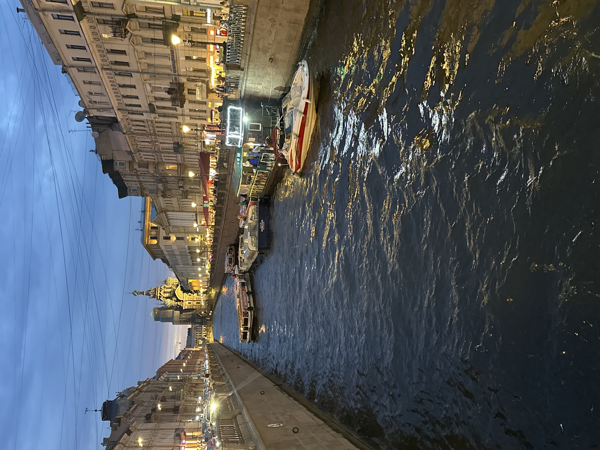 The image depicts a canal in Saint Petersburg, Russia, likely the Griboyedov Canal, as suggested by the presence of the Church of the Savior on Spilled Blood in the background. The scene is captured at dusk, with the sky displaying a deep blue hue transitioning into lighter shades towards the horizon. The canal is flanked by multi-story buildings, characterized by their ornate architecture and illuminated windows. Streetlights and lights from the buildings cast a warm glow on the water and the canal walls.
Several boats are docked or navigating the canal. In the foreground, a white and red speed boat is visible, its hull reflecting the ambient light. Behind it, a larger, covered tour boat with people on deck and illuminated by strings of lights is moored. Other boats, including a long, white vessel, are also present. The water is dark and rippled, reflecting the lights from the surrounding cityscape.
On the sidewalks lining the canal, numerous people can be seen walking and gathered. The buildings along the canal feature balconies and varied architectural details. Overhead, a network of power lines crisscrosses the sky. The overall atmosphere is that of a lively evening scene in a historic European city.