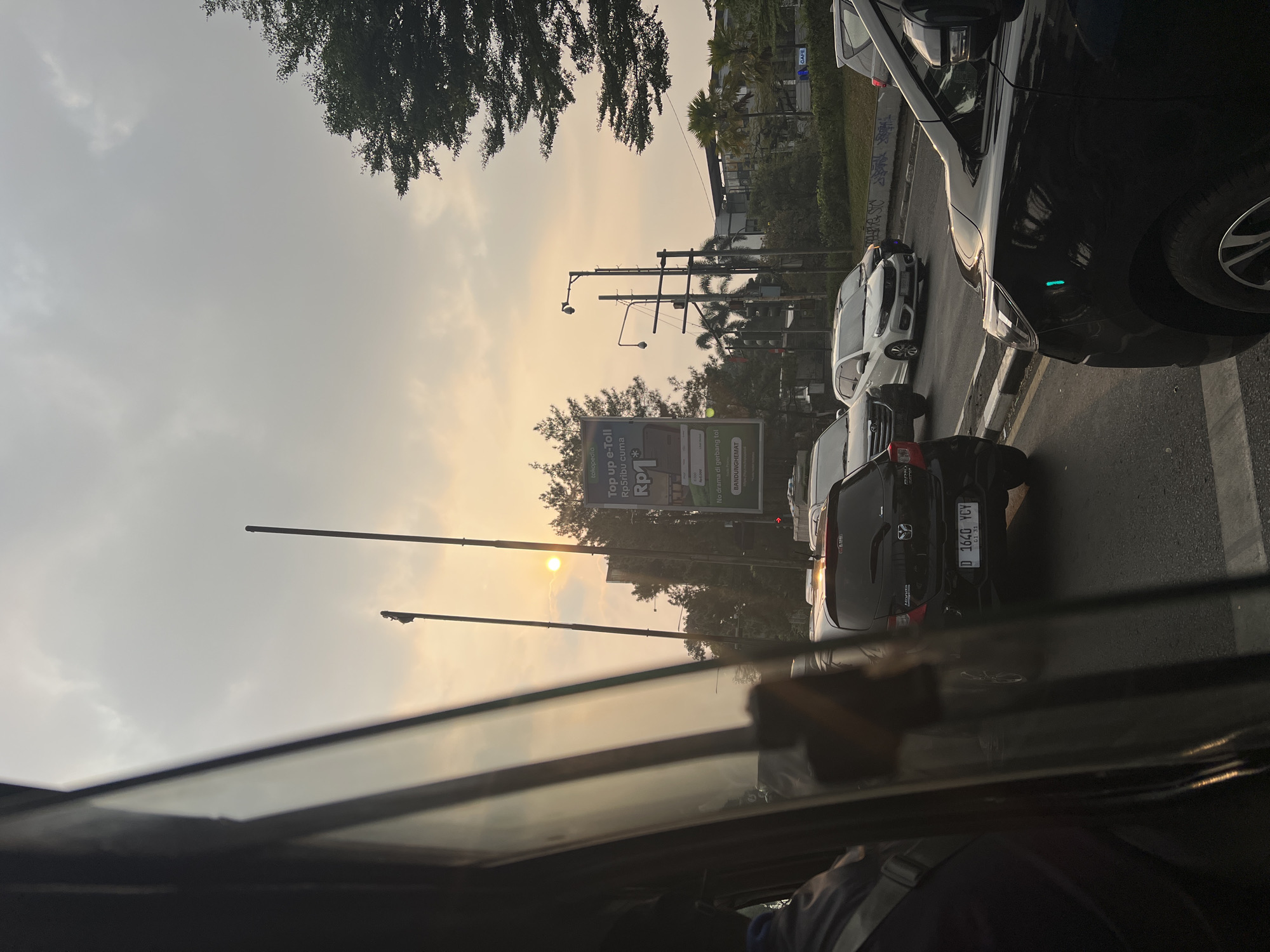 An outdoor, medium shot captures a sunset scene in Bandung City, Indonesia. The sun is visible as a bright orb in the sky, partially obscured by clouds. The sky is a hazy orange and gray. A traffic jam is evident, with several cars stopped on a paved road. Among the vehicles is a black hatchback with the license plate D 1640 YCY. A billboard advertising "Top Up e-Toll Rp1" is visible on the right side of the road, alongside palm trees and other vegetation. The image is taken from inside a vehicle, with the window reflecting the sky and other cars. The overall atmosphere suggests a typical urban evening commute.