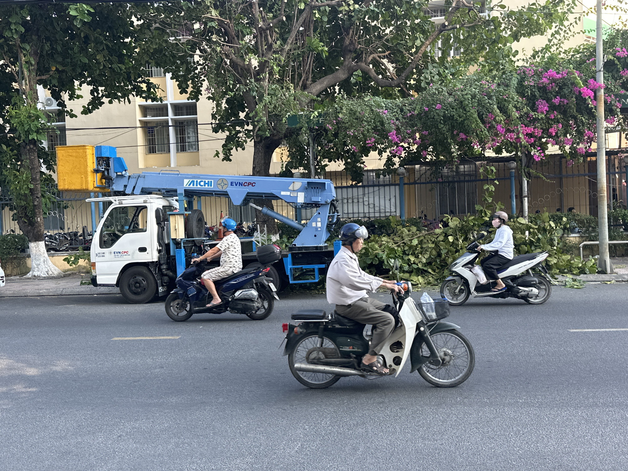 The image captures a street scene in the city of Nha Trang Ward, Vietnam. Three individuals are riding motorbikes on an asphalt road. The first individual, positioned centrally in the frame, is riding a green motorbike. The second individual, located to the left and slightly behind, is on a blue motorbike. The third individual, to the right and also slightly behind, is on a white motorbike and is wearing a face mask.
A white truck with a blue crane is parked on the side of the road. The truck has the text "AICHI" and "EVNCPC" on its side. Debris from tree trimming is scattered along the roadside. Trees are visible in the background, some of which have pink flowers. A building with multiple windows is partially visible behind the trees.