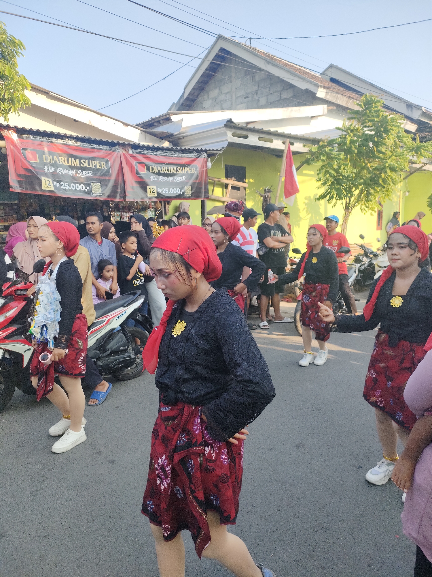 In Tongaswetan, Indonesia, a group of young women in red headscarves and traditional clothing participate in a parade. The women are adorned with flower pins on their black tops and wear red skirts with floral patterns. One woman in the foreground is decorated with small bills attached to her attire. Behind them, a crowd of onlookers watches the procession, some standing near motorcycles and a store displaying banners. The scene is set against a backdrop of buildings and trees under a clear sky.