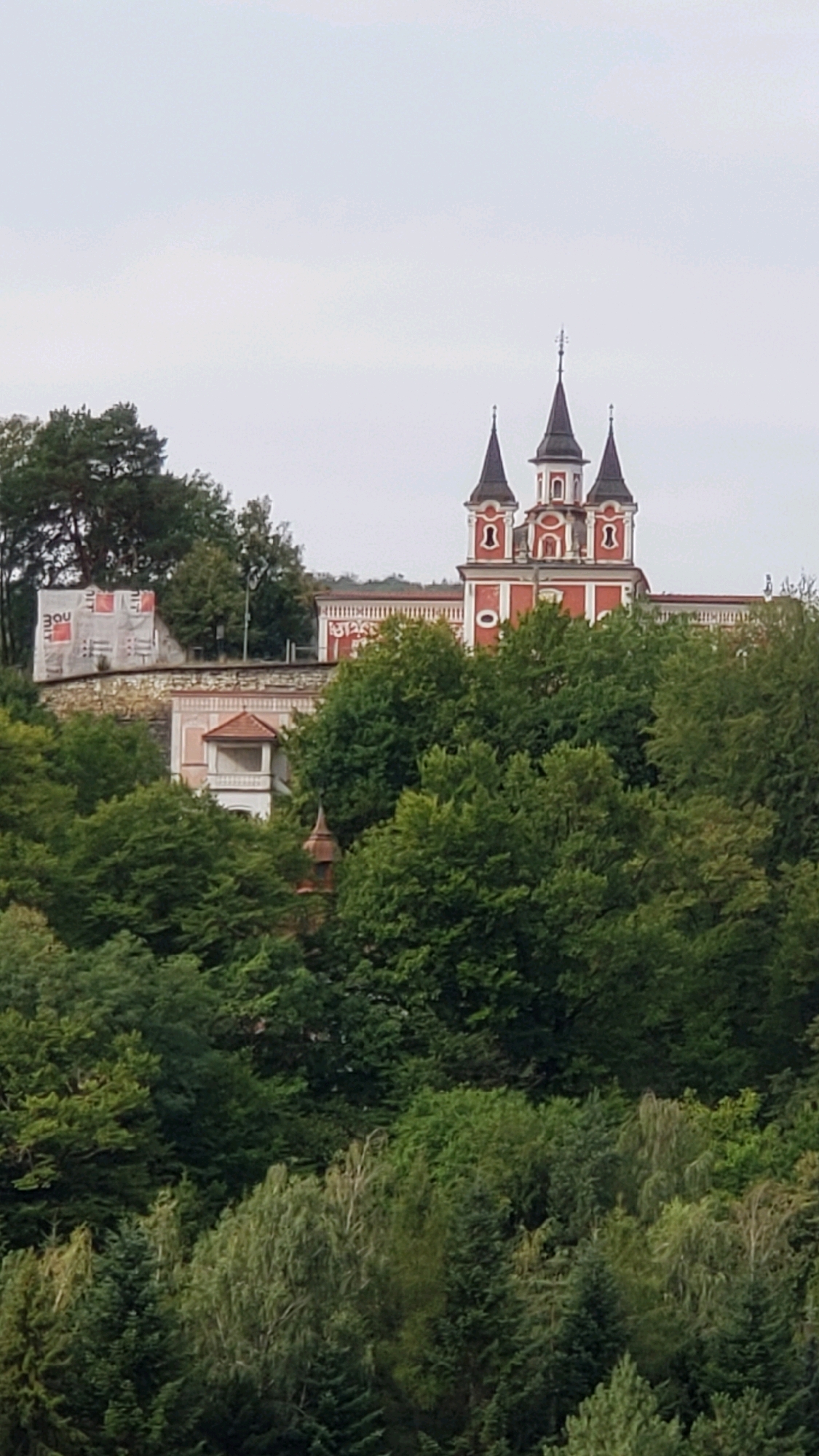 The image shows a view of the Orthodox Monastery of Saint John the Merciful (Monaster svätého Jána Milostivého) in Prešov, Slovakia, nestled amongst dense green trees. The monastery features a prominent red building with three dark, pointed spires, each topped with a cross. The architecture suggests a church or a significant religious structure. To the left of the main building, a lower section of the monastery complex is visible, with a light-colored wall and a small, enclosed balcony. Part of the wall is covered with scaffolding and white sheeting, indicating ongoing construction or renovation. The foreground and midground are dominated by a lush canopy of various shades of green trees, obscuring much of the lower parts of the monastery and suggesting a hilly or elevated location. The sky is overcast and pale. No people are visible in the image.