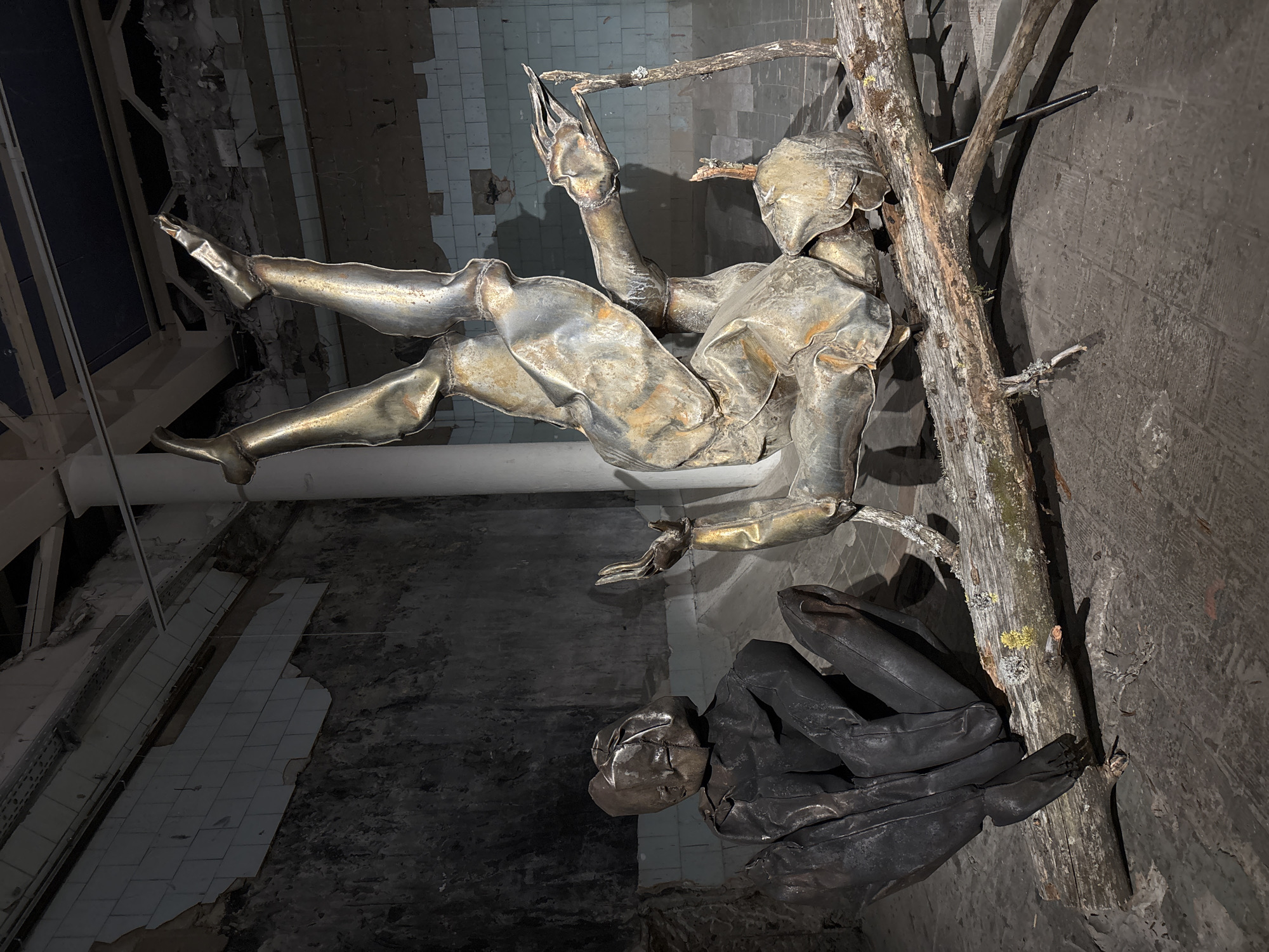 The photograph depicts a metal sculpture of a figure leaning against a tree branch inside a decaying building. The sculpture is constructed from crinkled sheets of metal, with one figure appearing to sit or kneel below the leaning figure. The environment is characterized by cracked tiles and decaying walls. The location is implied by the request to be in Saint Petersburg, Russia.