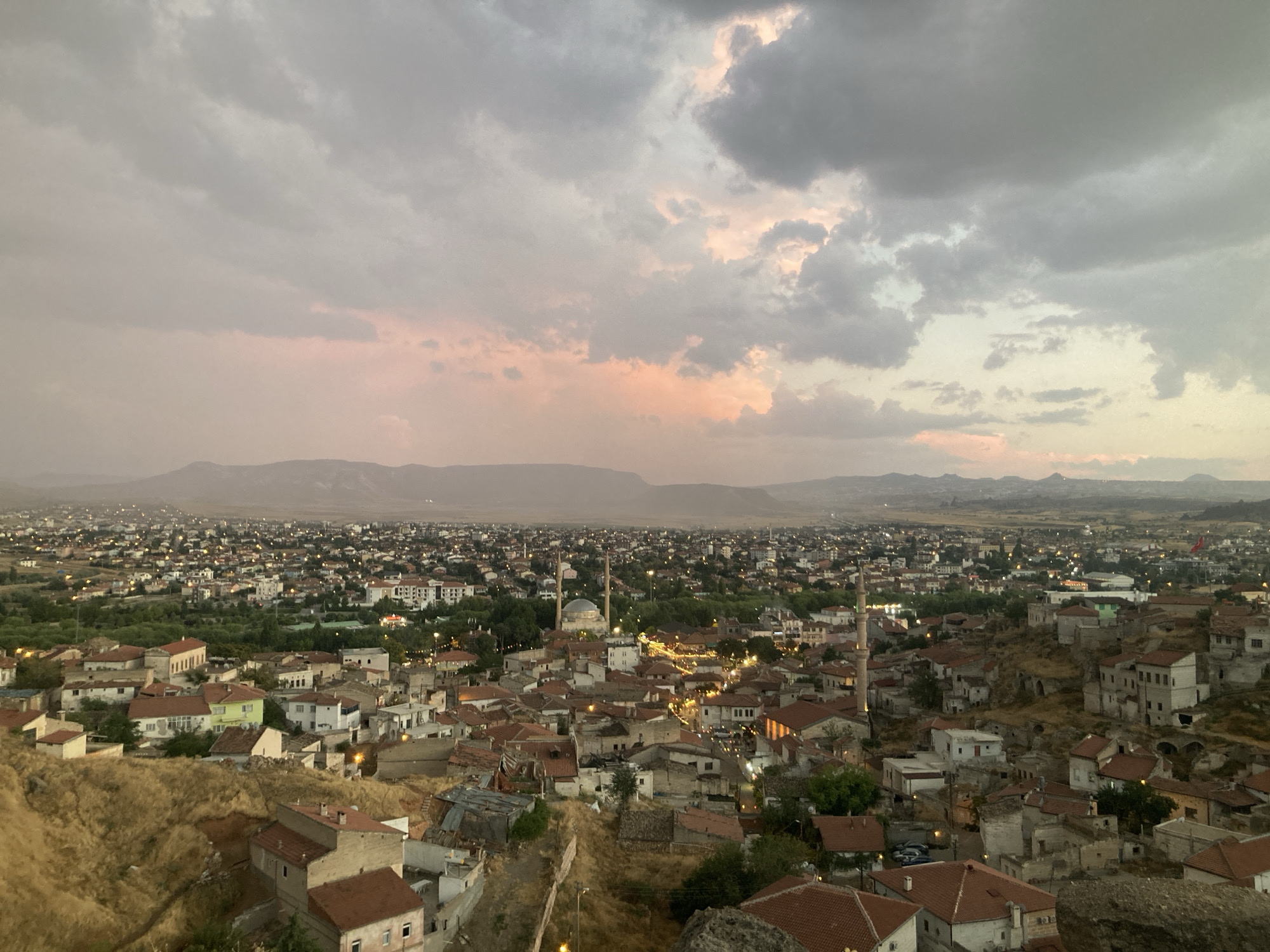 The image shows a panoramic view of Avanos İlçe Merkezi, Turkey, at dusk. The sky is filled with dramatic clouds, with soft hues of pink and orange indicating the setting sun. Below the sky, a sprawling city unfolds, characterized by a dense collection of buildings with red-tiled roofs, typical of the region.
In the foreground, a steep, dry, and rocky hillside is dotted with older, weathered structures, some appearing to be carved into the earth. As the eye moves further into the cityscape, newer buildings and a more organized urban layout become apparent. The city is illuminated by scattered lights, suggesting the transition from day to night.
Prominent architectural features include several minarets and domes, indicating the presence of mosques, most notably a larger one with a dome and two minarets in the mid-ground. The overall impression is of an ancient city with a rich history, blending traditional architecture with modern development, set against a natural, somewhat arid landscape. The mountains in the far distance are silhouetted against the twilight sky. There are no discernible people or active human interactions visible in the scene.