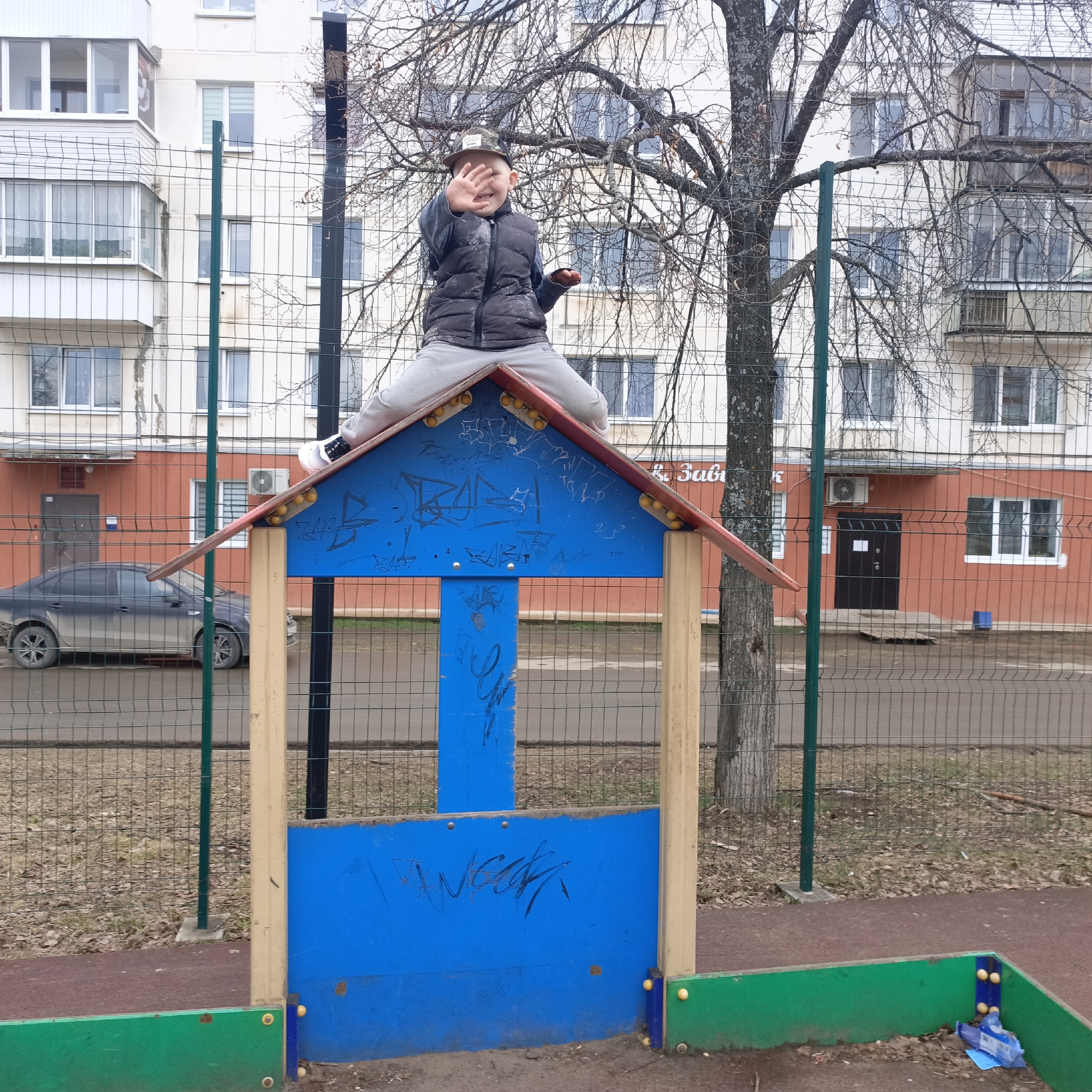 A boy is sitting on top of a blue, roofed structure in a playground. The structure is surrounded by a fence. In the background are apartment buildings, a parked car, and a tree with bare branches. The playground appears to be in a city, characterized by the urban architecture.