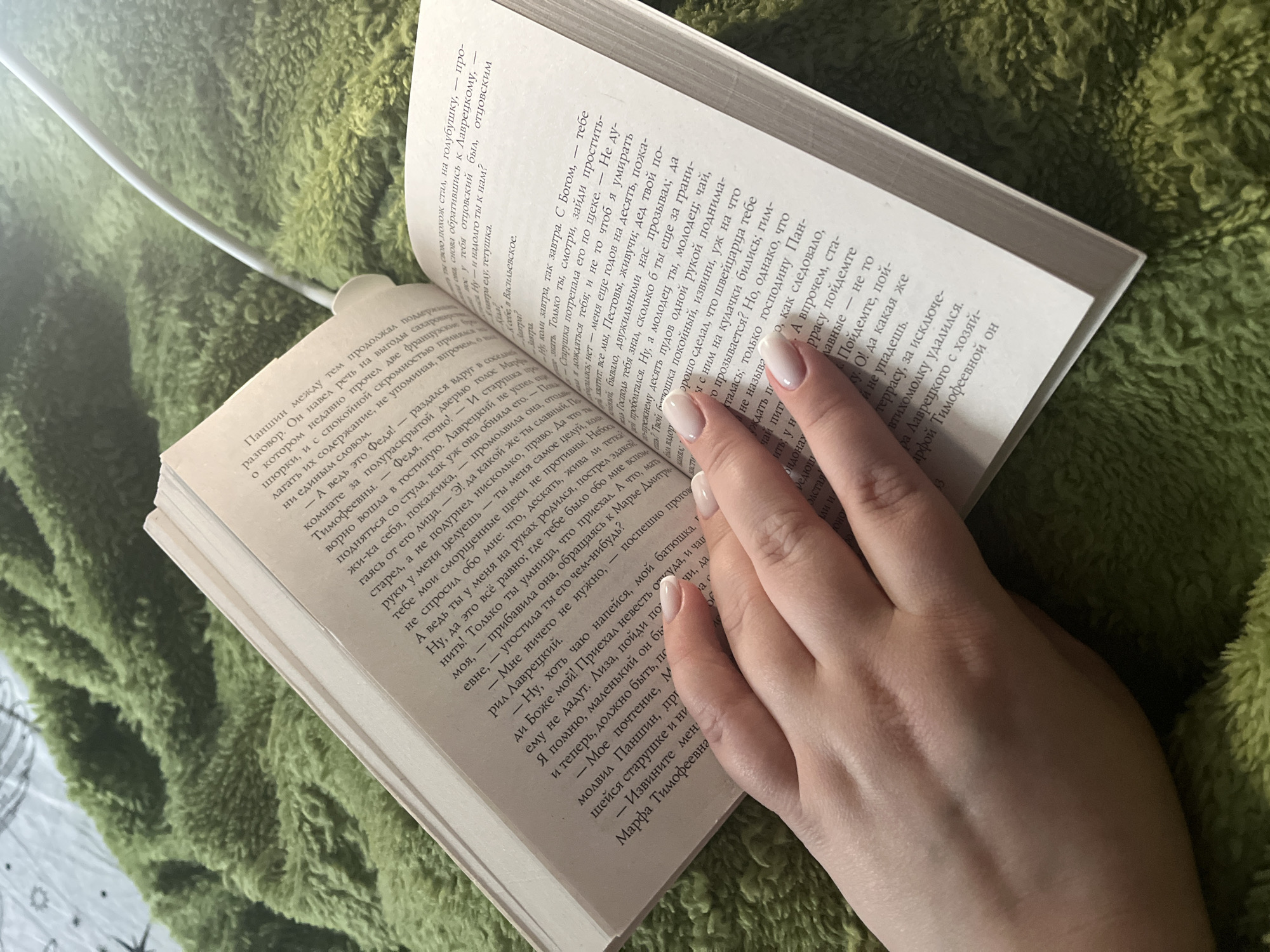 A book rests open on an unidentifiable green surface, a hand placed on the right page to hold it open. The book is written in Russian. A white cord or cable extends from the book's spine.