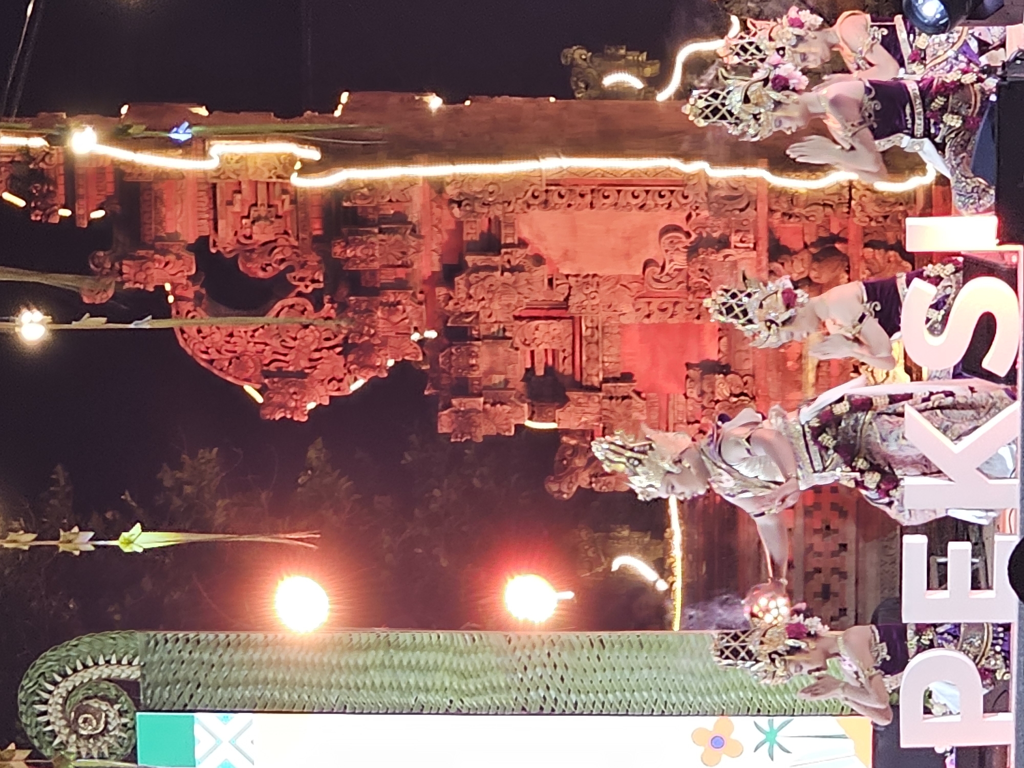 Captured at night in Denpasar, Indonesia, the image showcases Balinese dancers in ornate costumes and headdresses, kneeling in a line. The setting is adorned with traditional Balinese architectural elements decorated with string lights. The dancers are positioned in front of large white letters that are part of a sign or display. The foreground features a platform covered in woven material, with lights illuminating the scene. In the background, dense vegetation is visible, set against a dark night sky.