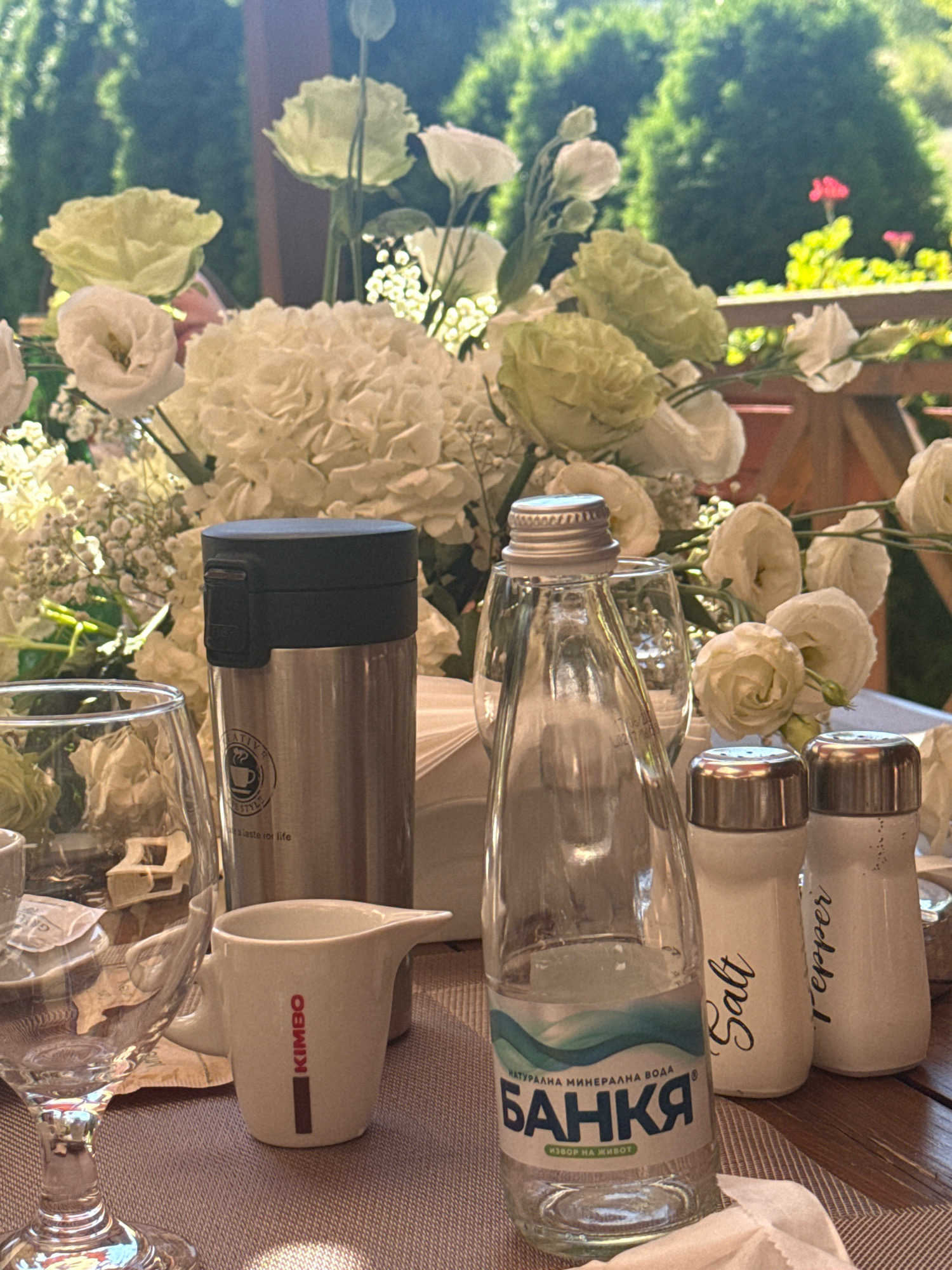 The image depicts an outdoor table setting with floral arrangements in the background. In the foreground, there are several objects on the table including a glass, a silver thermos, a small white pitcher with red lettering that reads "KIMBO," a clear glass bottle of water labeled "БАНКА," and two salt and pepper shakers. The bottle of water is of "NATURAL MINERAL WATER" from "BANKA" (meaning bank in Bulgarian), "SOURCE OF LIFE." The salt and pepper shakers are labeled "Salt" and "Pepper" in cursive. The background features lush green trees and a wooden railing. The lighting suggests it is daytime, possibly late morning or early afternoon, with sunlight filtering through the foliage. The presence of the "BANKA" mineral water suggests the location could be Bansko, Bulgaria, a town known for its natural springs and mineral water sources. There are no people visible in the image.