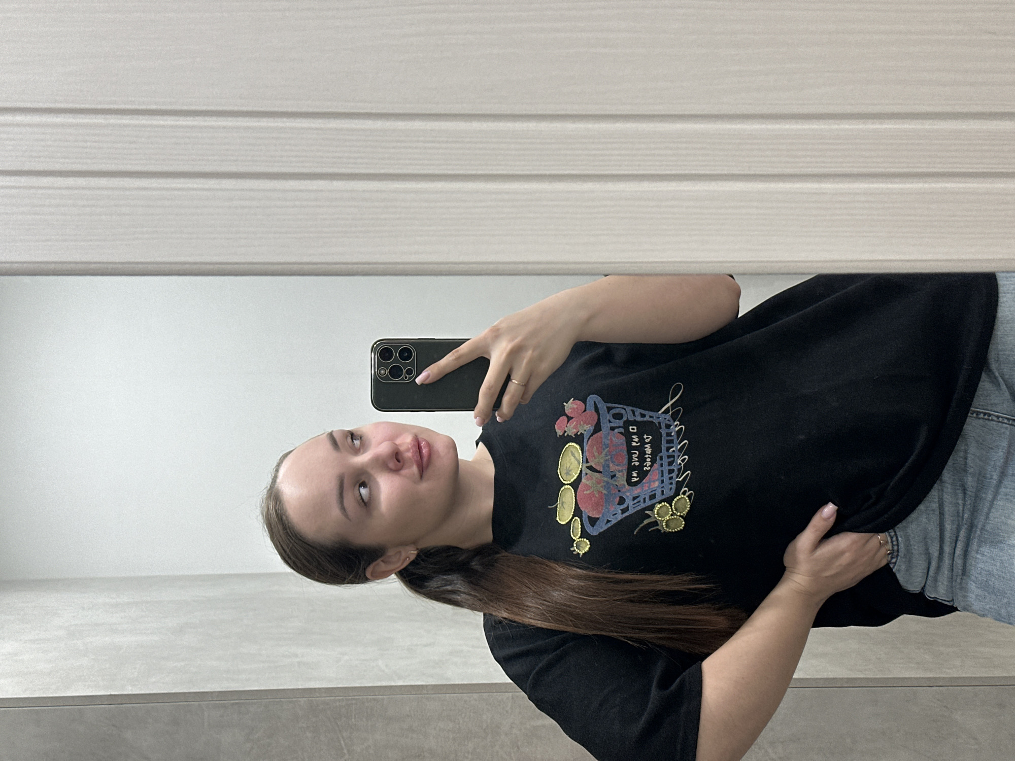 A young woman is taking a selfie in front of a mirror. She is wearing a black t-shirt with a design and light blue jeans. She has long brown hair pulled back in a ponytail. She is holding a black smartphone in her right hand to take the picture. The background includes a white paneled wall, potentially a bathroom. The location may be in the city of Chaikovsky, Russia, but there is no evidence of that in the image.