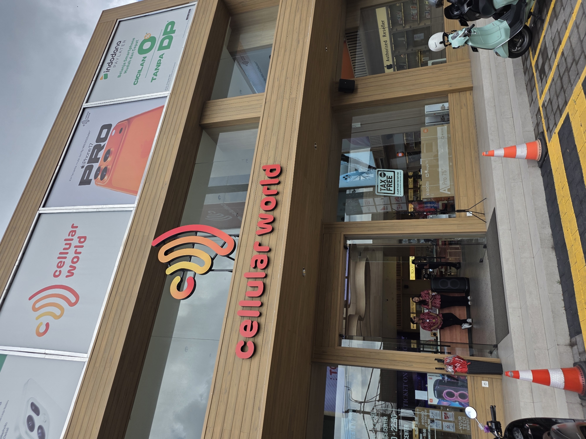 Here's a matter-of-fact description of the image:
The image depicts the facade of a "cellular world" store in a city (Unknown, Unknown). The store has a wooden exterior with large glass windows displaying promotional posters for smartphones (iPhone 17 Pro) and TV. There are also advertisement banners for Indodana Paylater with the tagline "Belanja Smartphone Mudah dan Cepat" and "Cicilan Tanpa DP". The store's name "cellular world" is displayed in large red letters.
The entrance is flanked by orange and white traffic cones. A light green scooter is parked near the cones on the right side. Through the glass entrance, two people can be seen inside the store, dressed in matching red shirts. There is also display for a JBL PartyBox. A "TAX FREE" sign is visible on the glass door. A black motorcycle is partially visible in the bottom right corner.