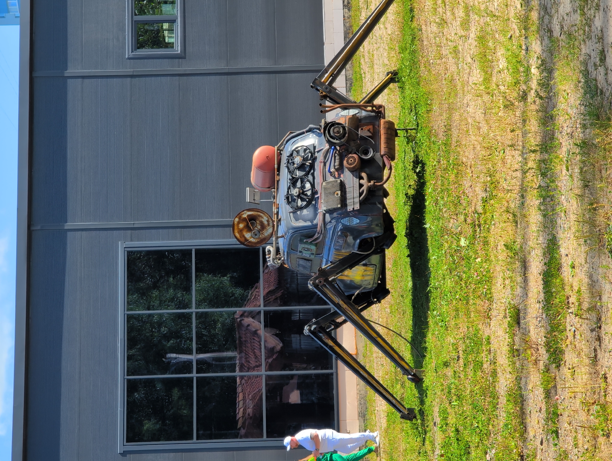 In Novoe Devyatkino, Russia, a bizarre, steampunk-inspired vehicle resembling a spider with a car body is displayed outdoors on a grassy area. The vehicle features multiple metallic legs that lift the car body several feet off the ground. The car itself appears to be an older model, modified with various mechanical parts, including what looks like an engine protruding from the front, fans, and a cylindrical tank on top. Two individuals are visible in the background, one wearing a white cap and shirt with white pants, and another in a green outfit. They are standing near a large, dark gray building with expansive windows reflecting the surrounding trees and rooftops. The environment suggests an outdoor exhibition or event space.