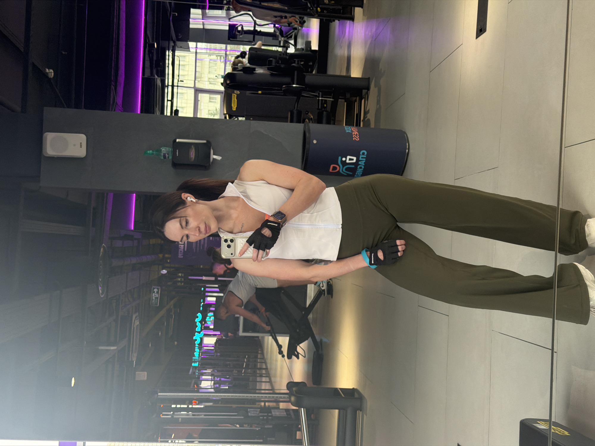 A woman is taking a selfie in a gym in Perm, Russia. The woman is wearing a white tank top and olive-colored pants. She is wearing workout gloves on her hands. She is holding her phone in her right hand. The gym has various workout machines. A person is working out in the background. The walls are gray and the floor is tiled. There is a sign with blue lettering visible. The gym is lit with purple neon lights.