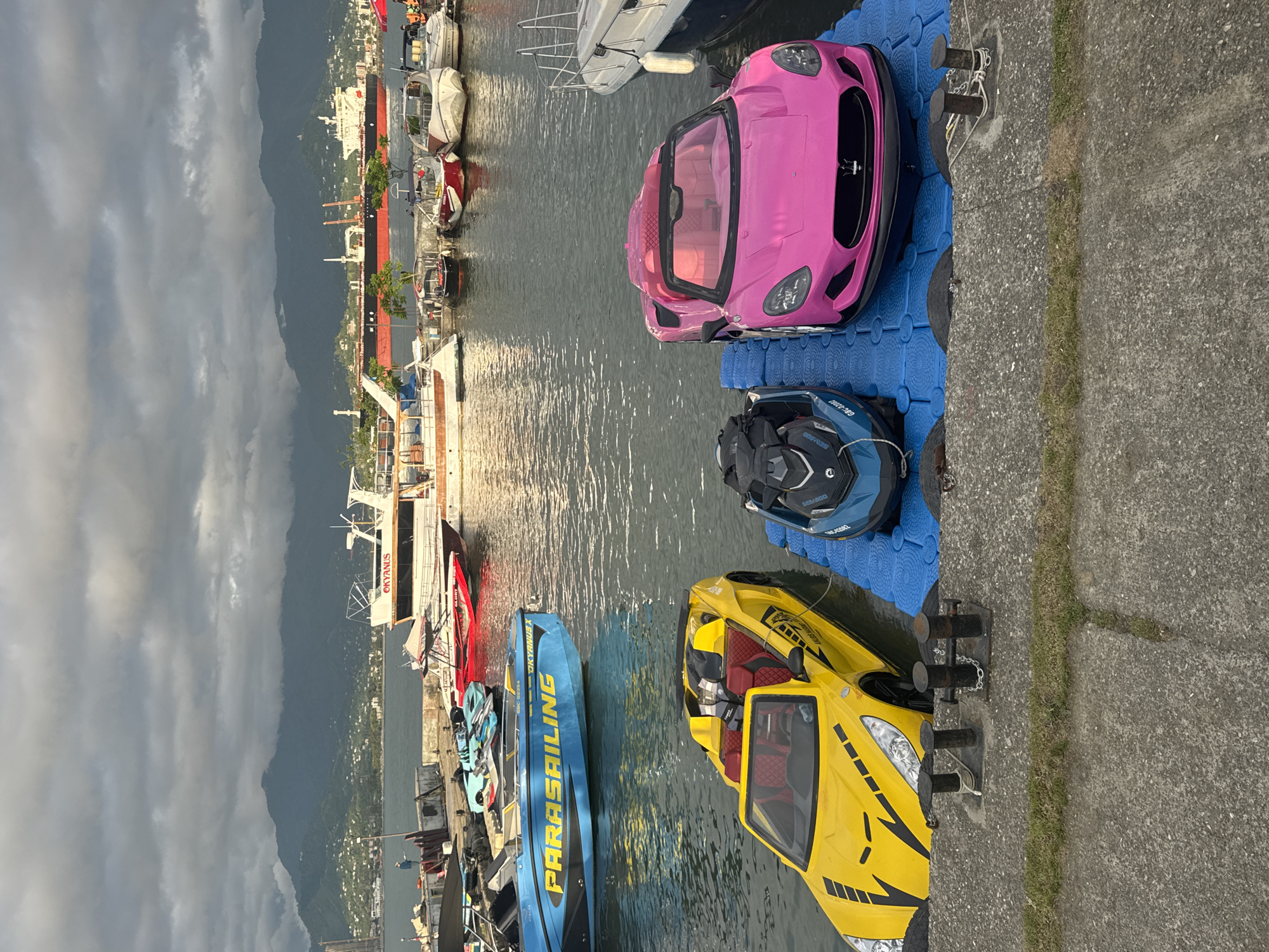 The image shows a harbor scene in Batumi, Georgia. In the foreground are a bright pink pedal boat shaped like a car, a blue jet ski, a yellow pedal boat shaped like a car, and a blue parasailing boat docked on a blue pontoon. Beyond the pontoon is a concrete embankment. The water reflects sunlight and sky. In the background are boats and buildings along the shoreline, and a distant mountain range is partially obscured by a cloudy sky.