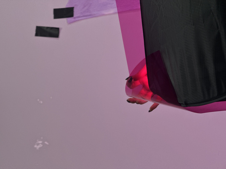 The image displays a human hand with dark nail polish, fingers extended, positioned behind a translucent magenta sheet that is internally illuminated, casting a prominent red glow around the digits. This foreground arrangement is set against a uniform light purplish-pink background. Below the illuminated magenta sheet, a section of dark, textured fabric, possibly nylon, with a subtle diamond-grid pattern and a horizontal seam, is visible. The upper left of the background contains several indistinct white speckles. In the upper right, two rectangular black adhesive strips are affixed to the background, beneath which a piece of purple fabric or paper hangs partially visible. No specific architectural or geographical features are present to discern a particular location within Sofia, Bulgaria.