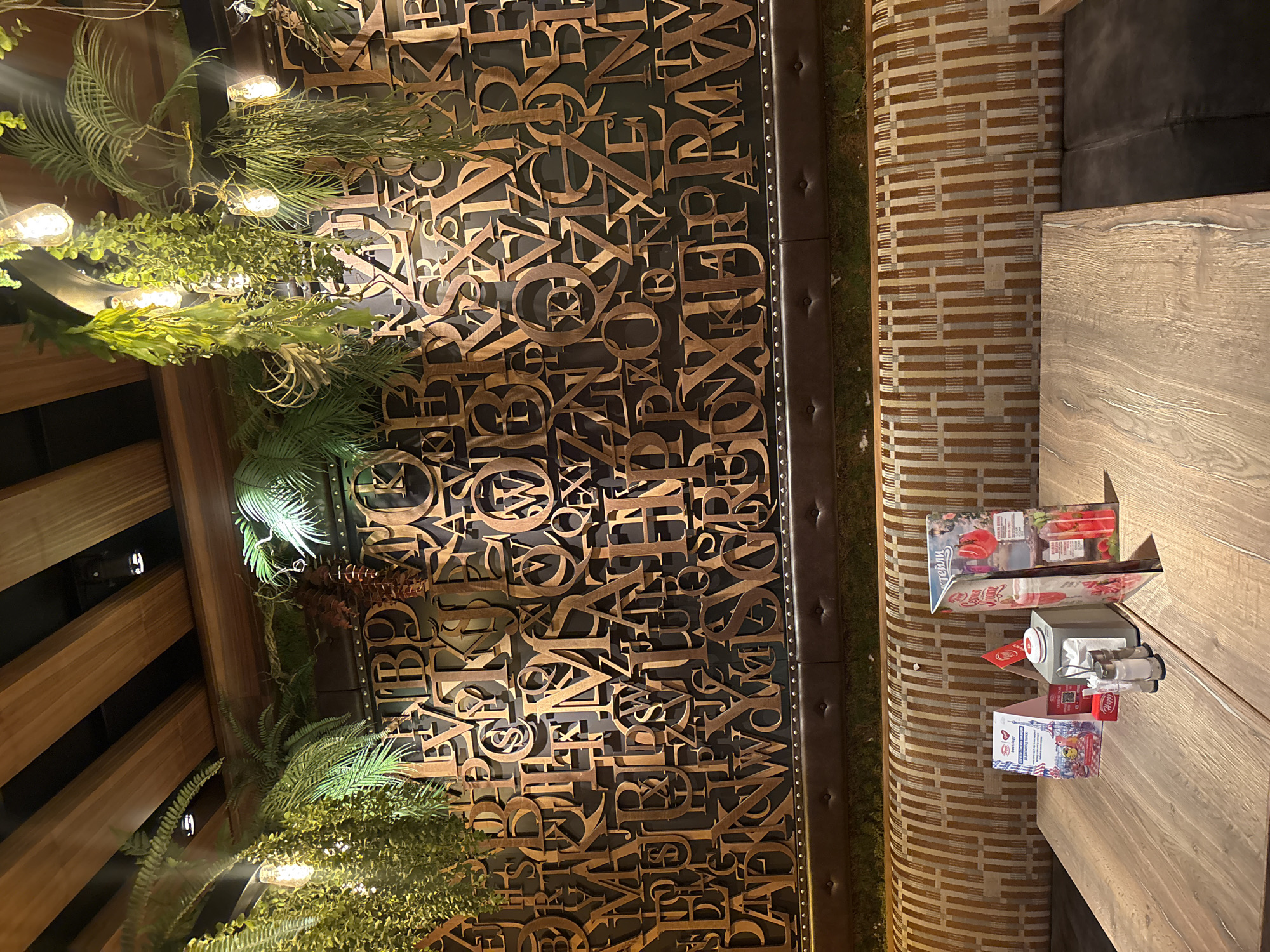 The image shows the interior of a restaurant or cafe in Plovdiv, Bulgaria. A decorative wall is covered in an arrangement of wooden letters. Green plants and lights adorn the wall on one side. A table with condiments and menus sits beside a patterned banquette. The table is wooden, and the banquette is upholstered in a patterned fabric. No people are visible.