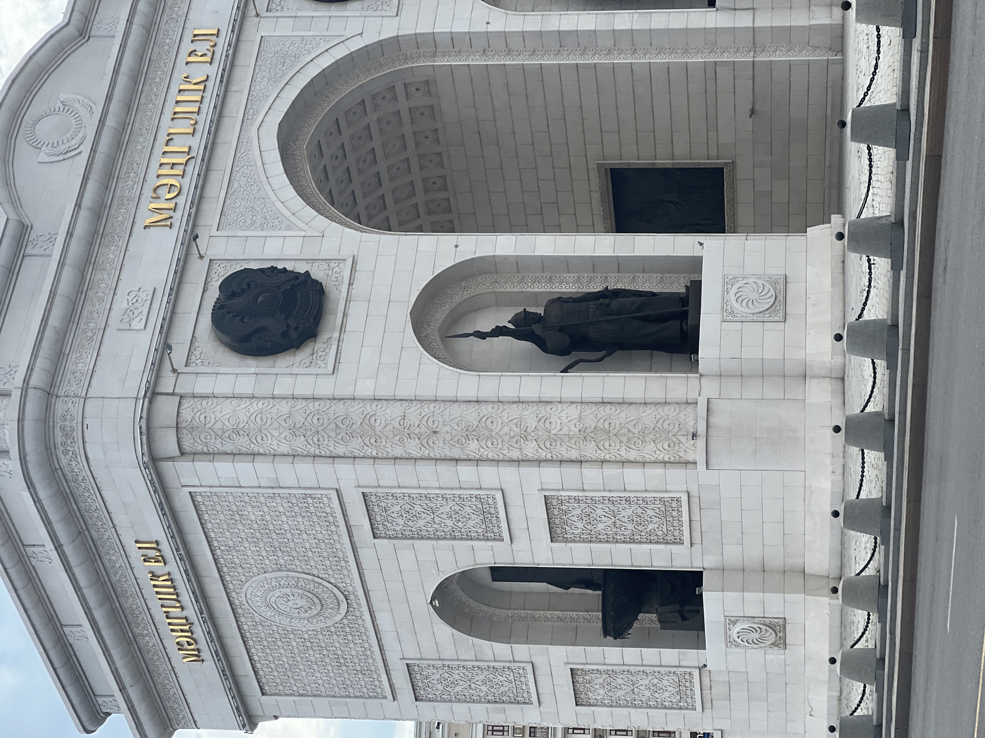 The image shows the "Mangilik El" (Eternal Nation) monument in Astana, Kazakhstan. The monument is a large, white, triumphal arch made of stone. It features ornate carvings and inscriptions in gold Cyrillic letters. There are two statues of figures in traditional attire holding spears, positioned within arched alcoves on either side of the main archway. The monument is situated in an urban environment, with a street and pavement in the foreground, and a low concrete barrier with chains separating the monument from the street. The sky is overcast.