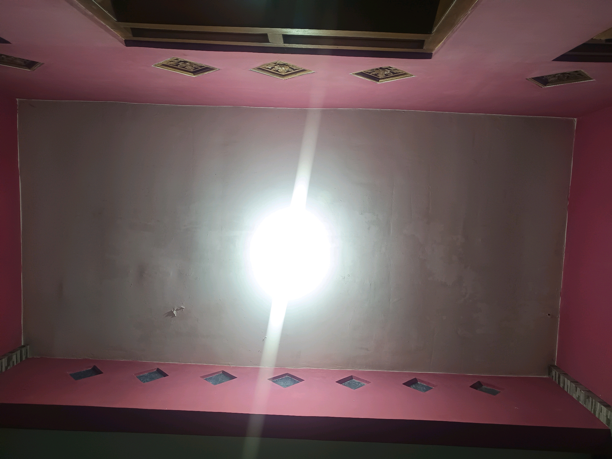 The image shows a low-angle view of a ceiling in a room. The ceiling is painted pink and features several decorative diamond-shaped inlays with gold patterns. A bright light source is centrally located on the ceiling, casting a strong glare. Below the main ceiling, there is a lower horizontal band, also painted pink, with several diamond-shaped cutouts. The walls are a darker pink color. The room appears to be indoors and likely residential. The location is identified as Gunung Sekar, Indonesia.