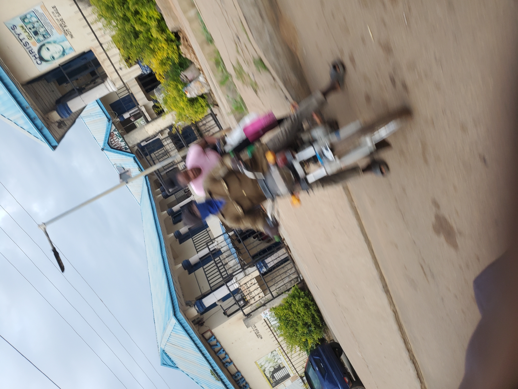 The image, taken in Bukuru, Nigeria, depicts a street scene with a two-story building featuring a light blue corrugated metal roof and white columns. A sign on the building reads "MARIGOLD PLAZA." Another sign, partially visible, advertises "CHRISTY'S GLAM." In the foreground, two individuals are on a motorcycle. The driver, wearing a brown jacket and a blue face mask, is partially obscured. A passenger, wearing a pink t-shirt, is seated behind the driver. The motorcycle is silver with visible wheels and a headlight. The street is paved with concrete, and there is vegetation on the right side of the image, along with some debris. A blue car is parked on the left side of the road. The overall image is blurry, suggesting movement.